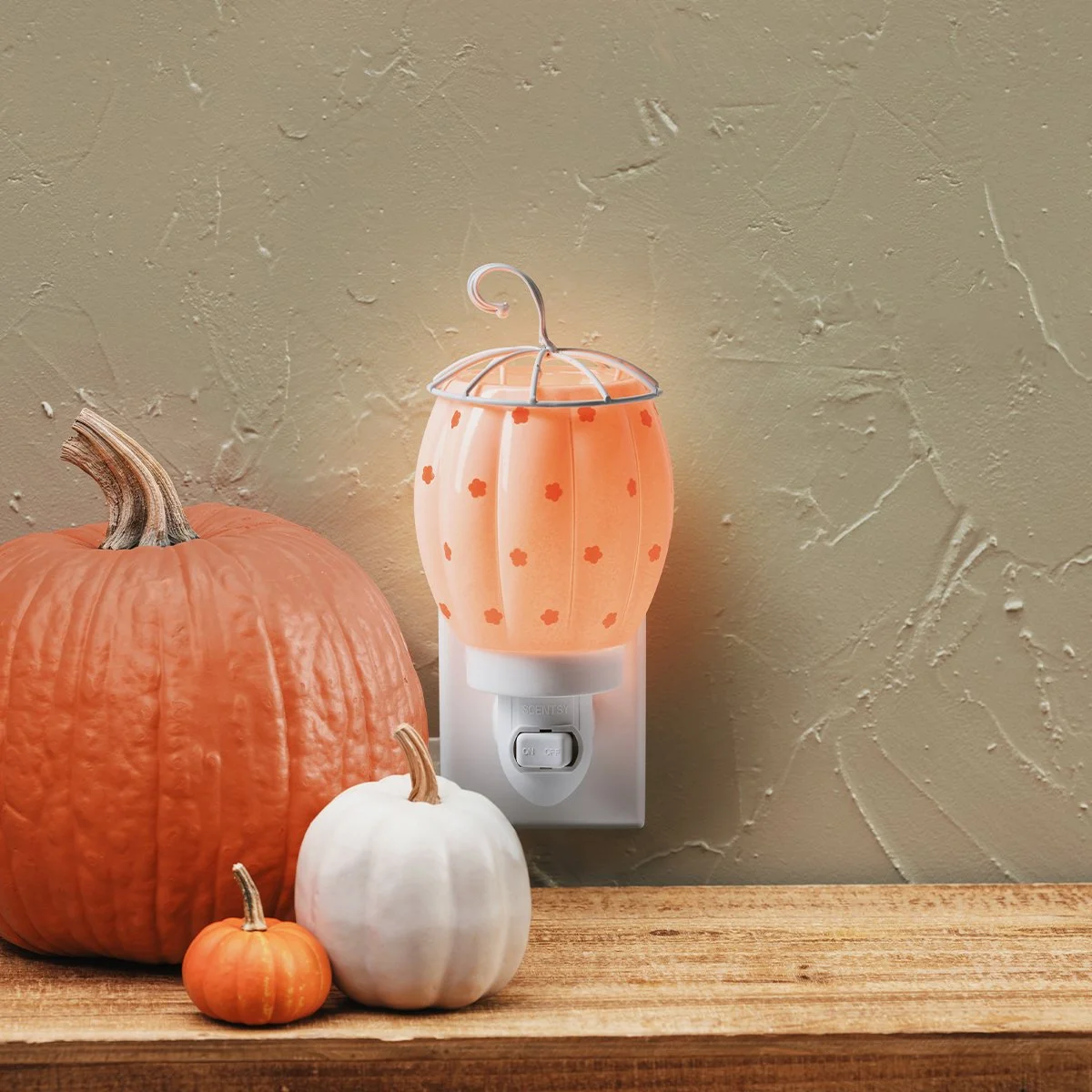 Scentsy Pumpkin Posy Mini Warmer glowing beside pumpkins on a wooden shelf, adding a soft autumn glow