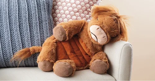 Brown Scentsy plush Highland cow sitting on a sofa with decorative pillows