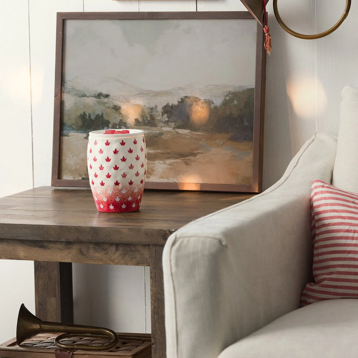 The Simply Maple Scentsy Warmer showcases a white ceramic finish decorated with red maple leaves, celebrating the timeless beauty of Canada. Displayed in a warm, rustic setting, this exclusive warmer brings a cozy and welcoming touch to any home