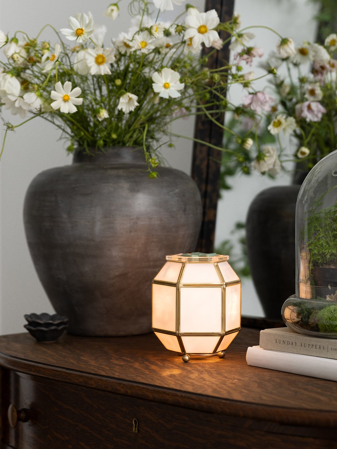 A softly lit Scentsy warmer with frosted glass panels and warm brass detailing styled on a vintage console with florals and neutral accents.