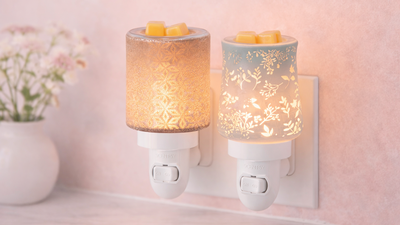 Two Scentsy mini wax warmers plugged into a wall outlet, featuring softly glowing glass and ceramic designs with a light, neutral background and warm ambient lighting.