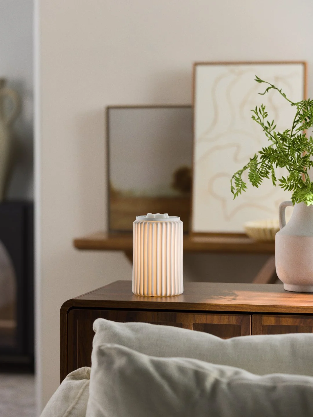 A white fluted Scentsy warmer with a gentle glow styled on a wood cabinet with neutral artwork and soft greenery for a calming, minimalist feel.