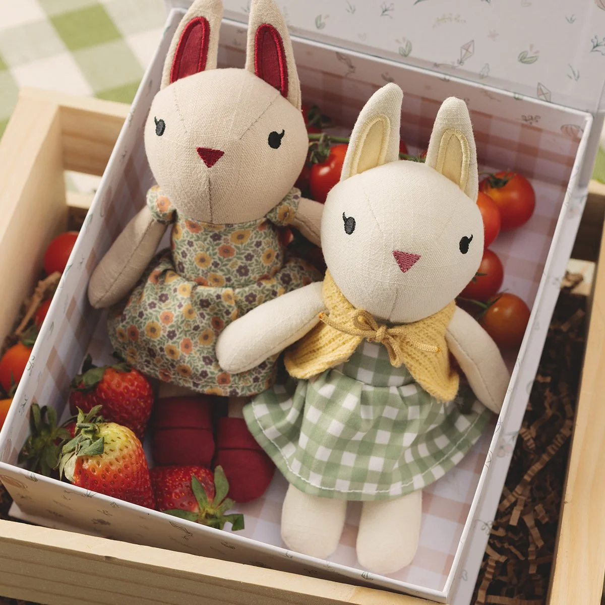 Two soft bunny plush figures from the Fresh-picked Friends collection sit in a crate with strawberries and tomatoes, styled to reflect a cozy farmers market theme.