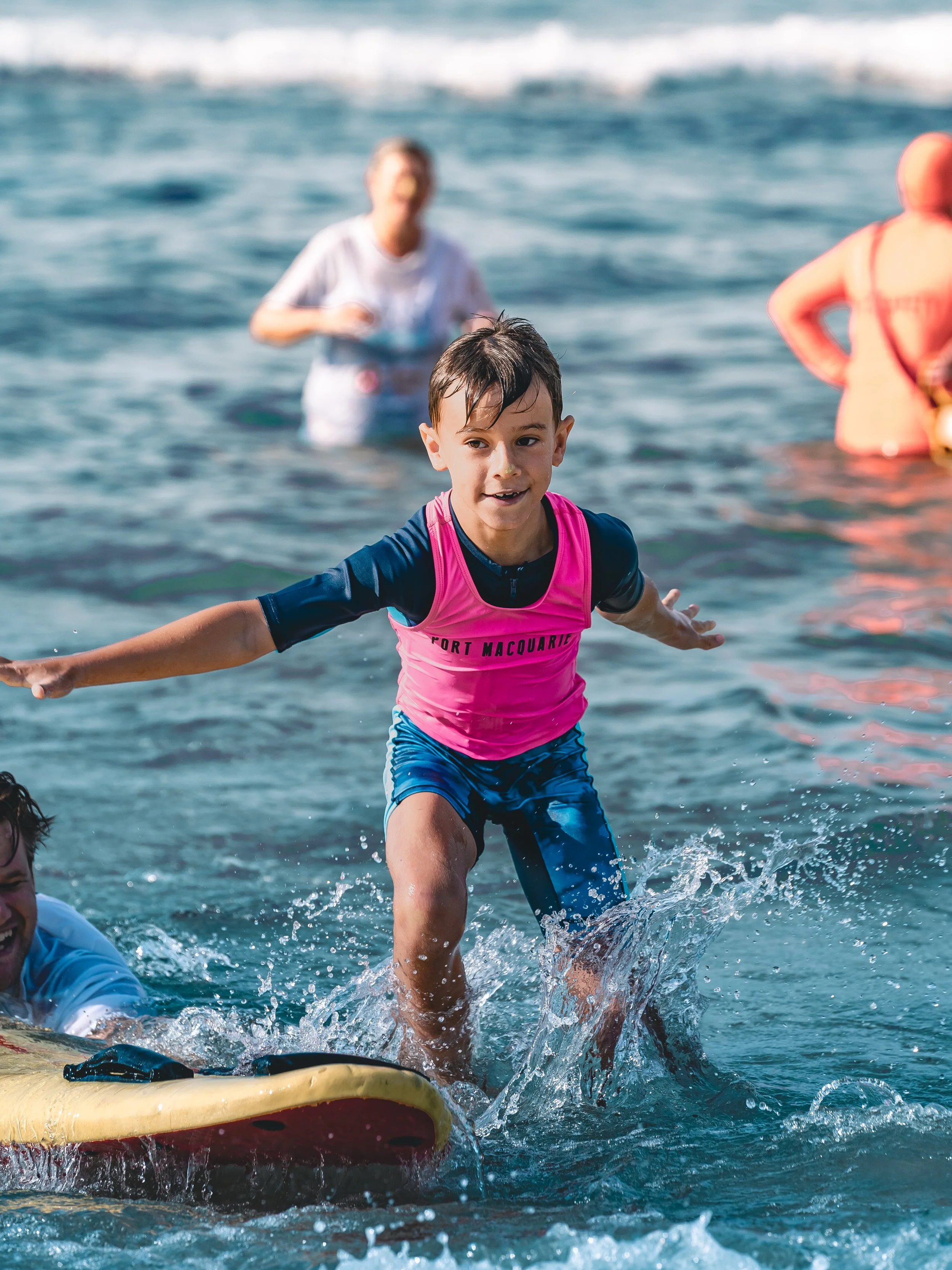 A young boy in a pink life jacket balancing on a surfboard in the water, with adults in the background, enjoying a day at the beach.