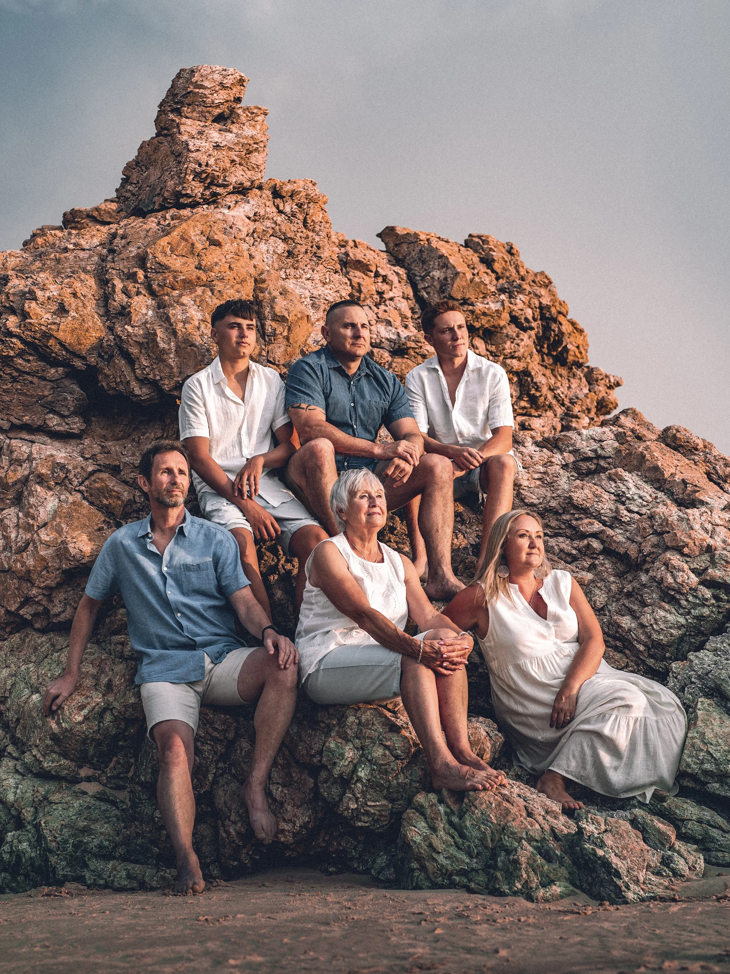 Seven people sitting and standing on rocks at the beach during sunset, looking towards the distance.