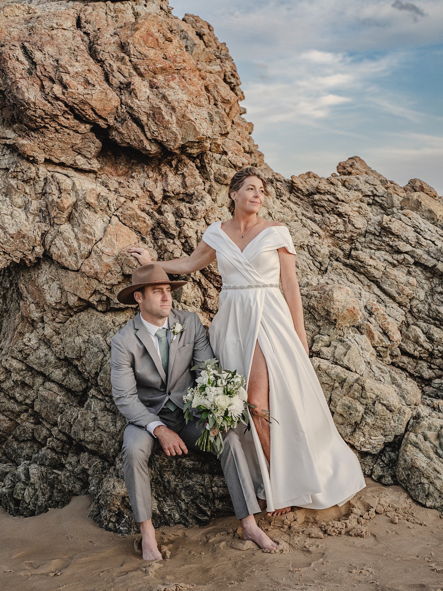 A woman in a white wedding dress standing on a beach next to a man in a gray suit sitting on rocks, holding a bouquet of white flowers. The woman is touching the rocks with one hand and looking away, the man looks thoughtful. The scene is outdoors with a rocky background and a partly cloudy sky.