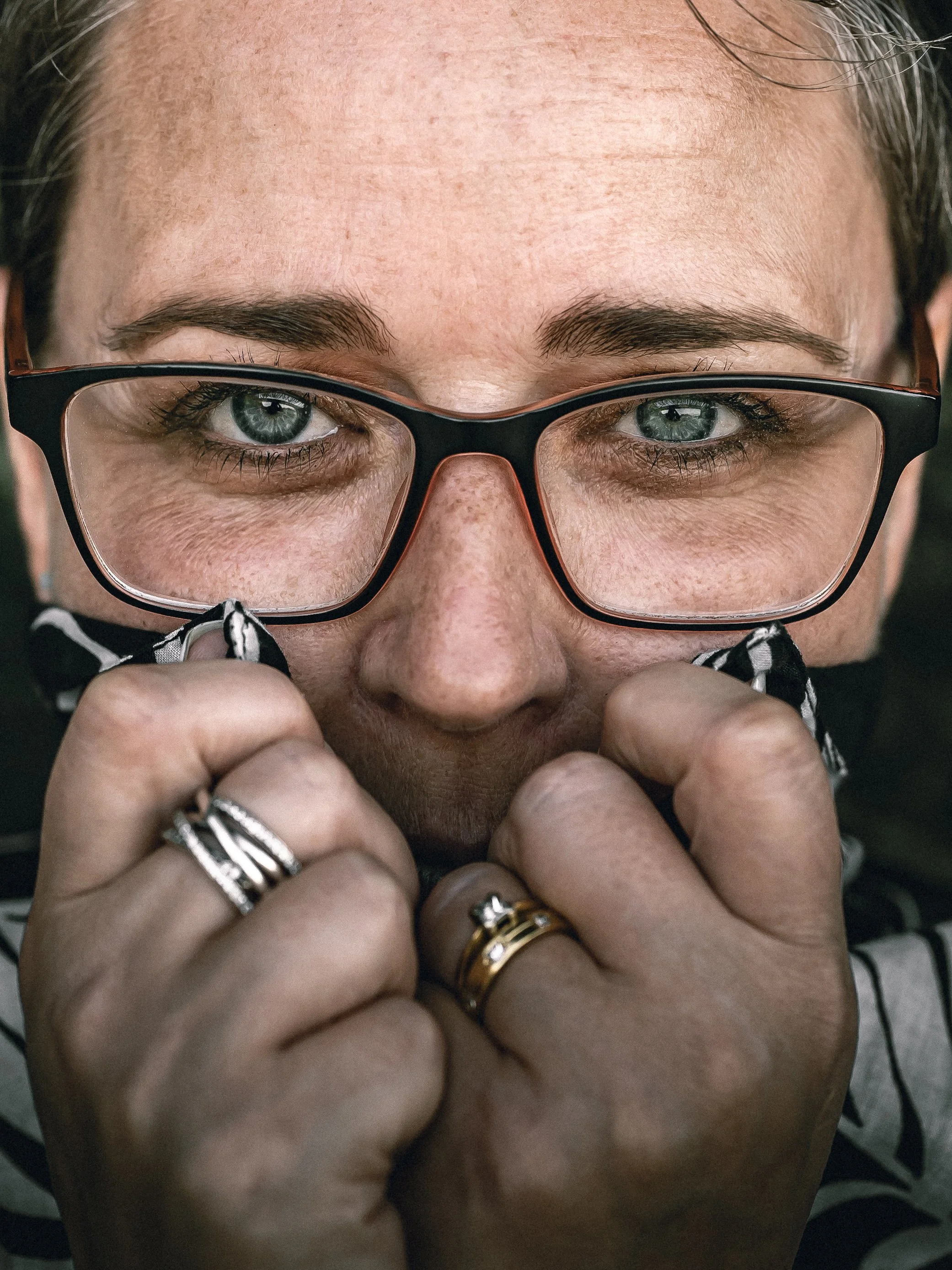 A close-up portrait of a woman with glasses, brown hair, and rings on her fingers, holding her glasses with both hands near her face, looking directly at the camera.