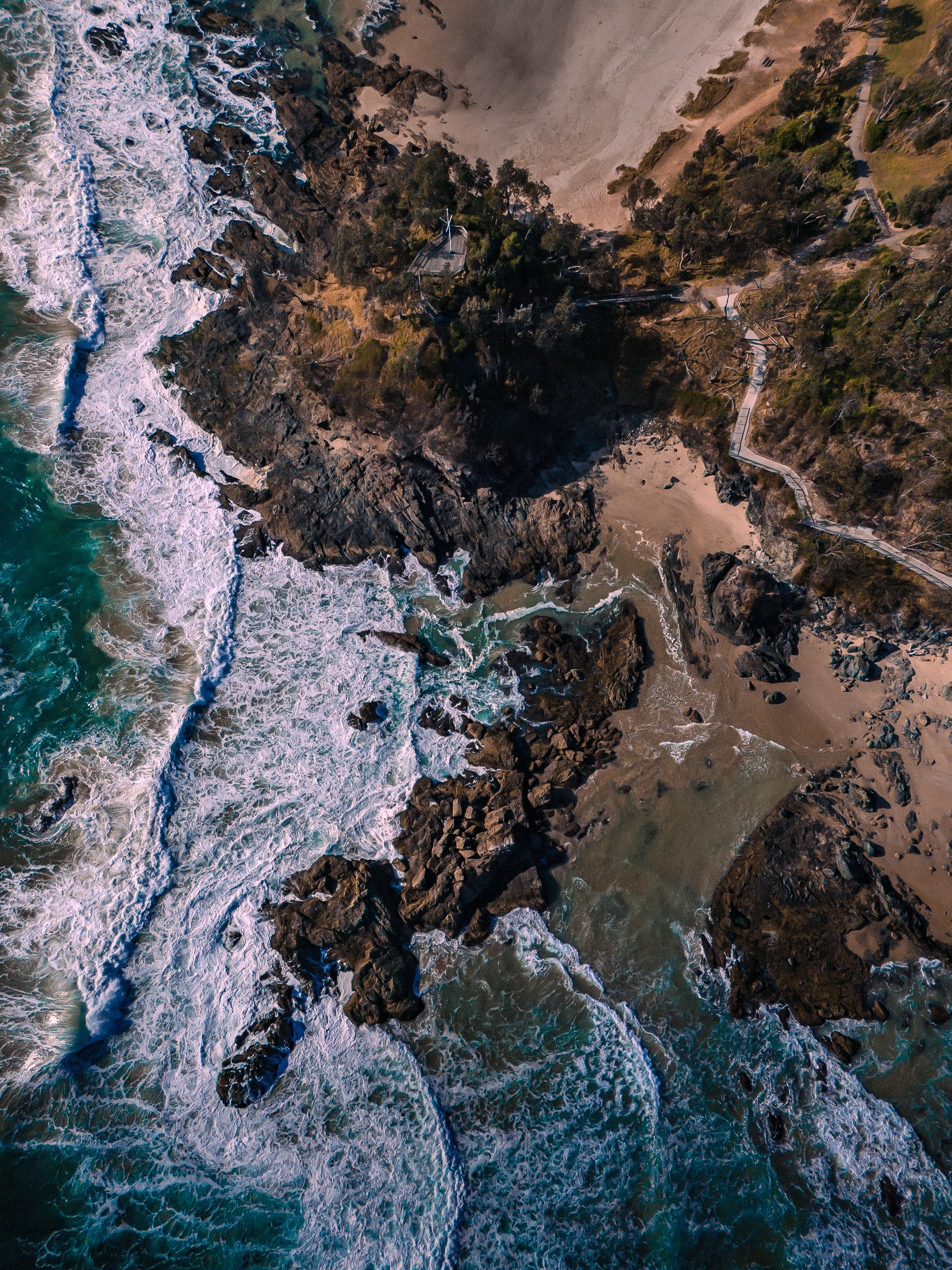 Aerial view of a rugged, rocky coastline with waves crashing onto the shore, a sandy beach area, and a green forested area with pathways visible.