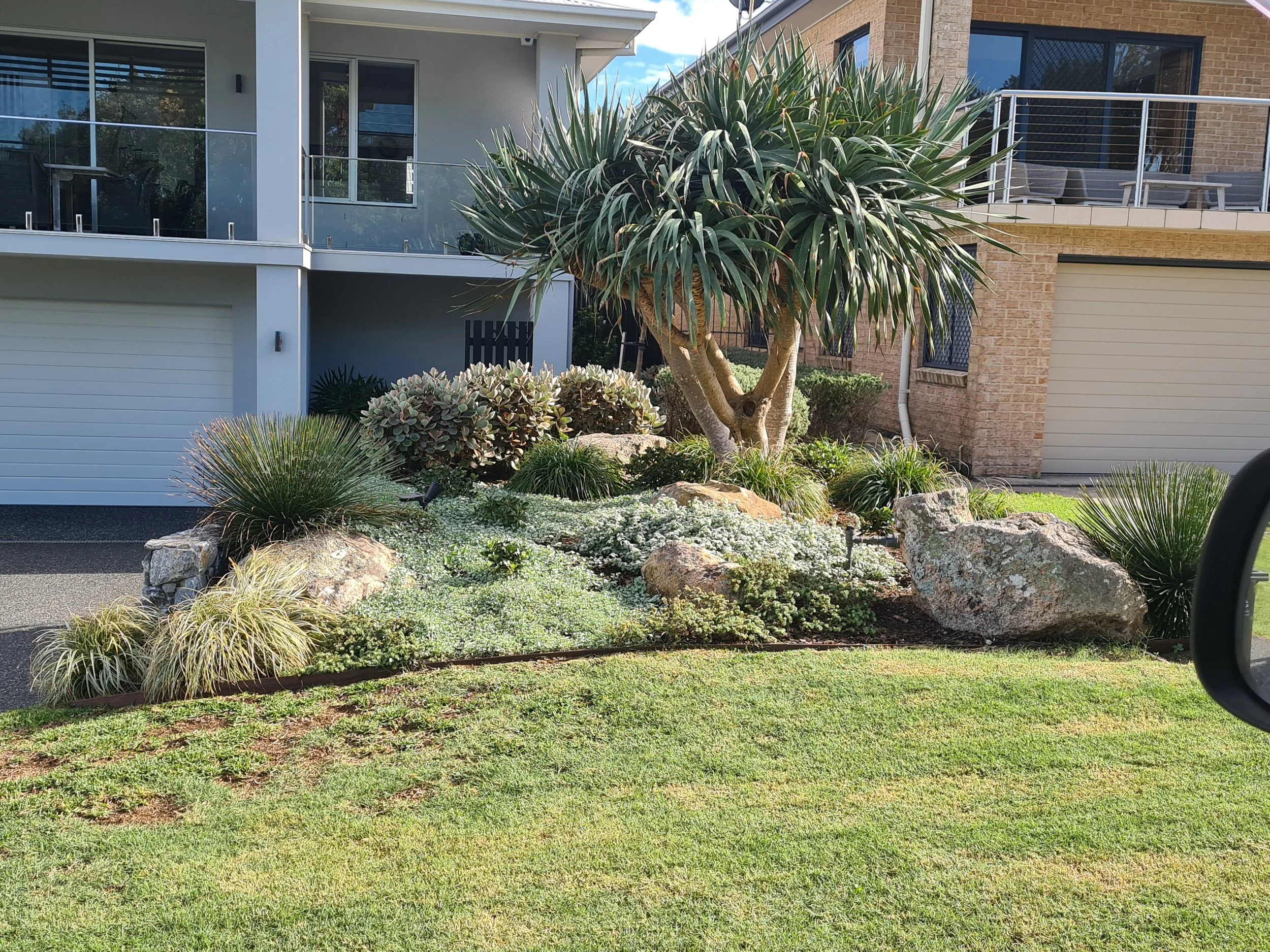 Modern house exterior with landscaped garden, featuring a variety of plants, rocks, and a grassy lawn. The house includes a garage and an upper balcony with glass railing.