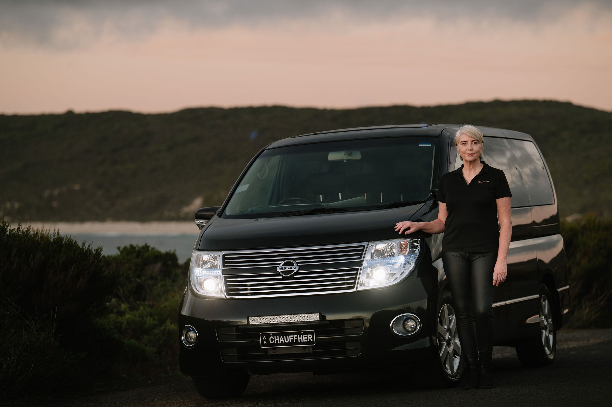 An image of Tracie standing beside the luxurious ElGrand chauffeur vehicle with Smiths Beach in the backrgound