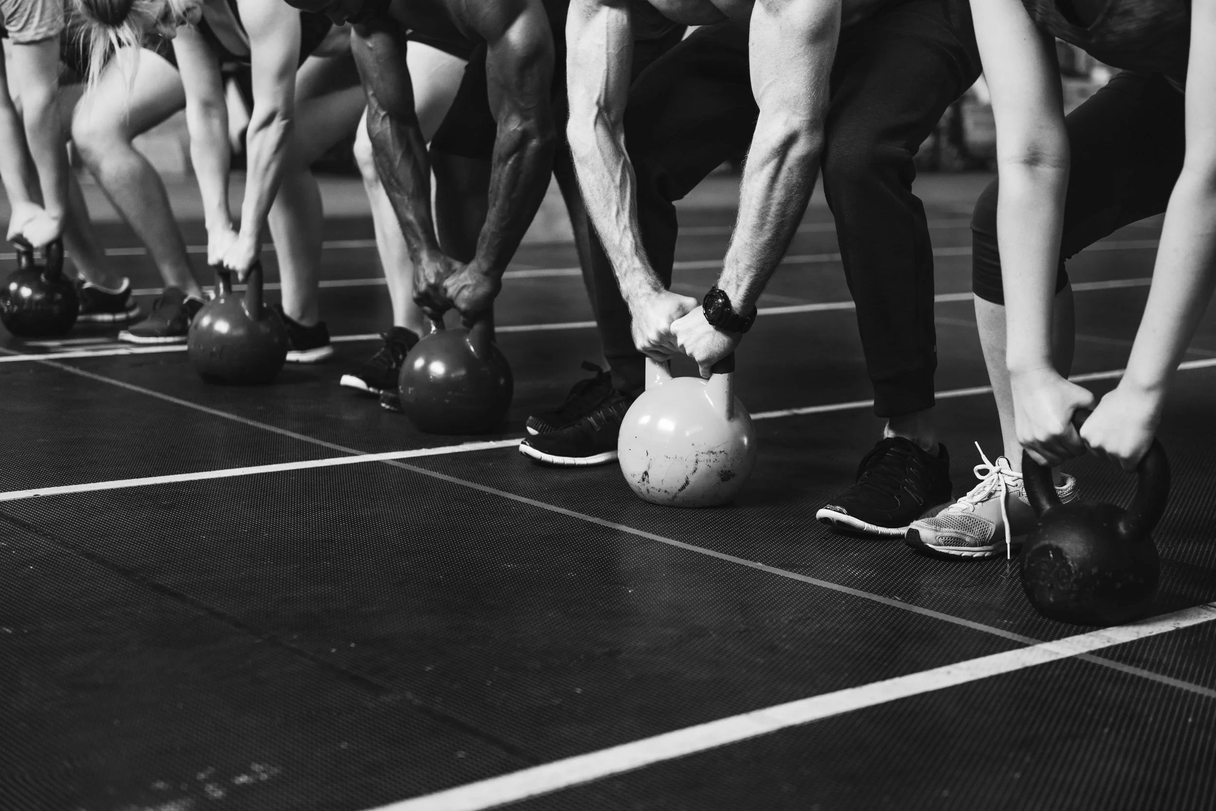 Group of people participating in a fitness class, performing kettlebell exercises on a gym floor, with a focus on their hands gripping kettlebells.
