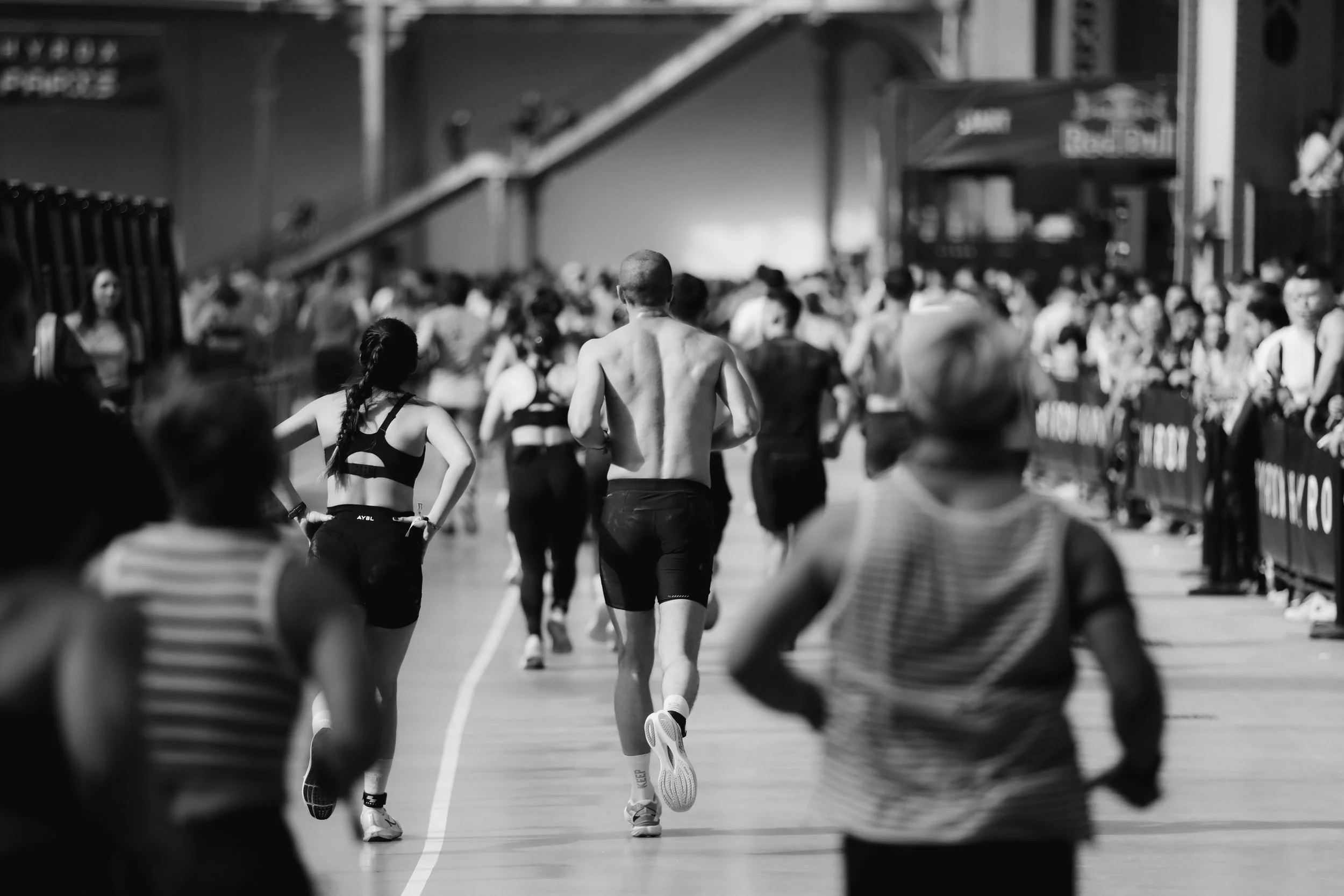 A black and white photo of a crowded indoor running track with people running and walking, with some spectators blurred in the foreground.