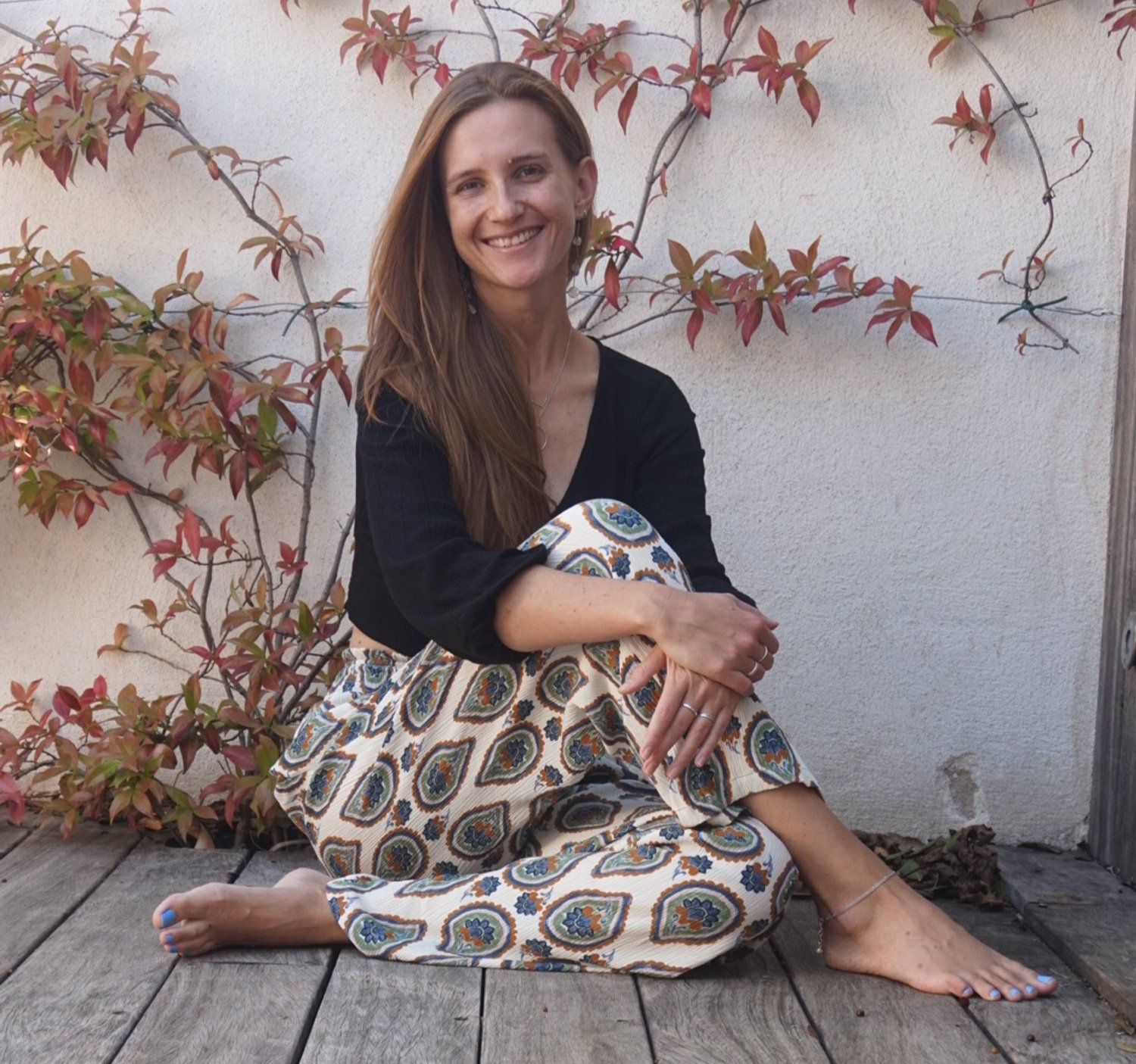 A lady (Alice) sits cross-legged on a wooden deck, wearing a black top and patterned pants, smiling at the camera. The background features a wall with climbing plants.