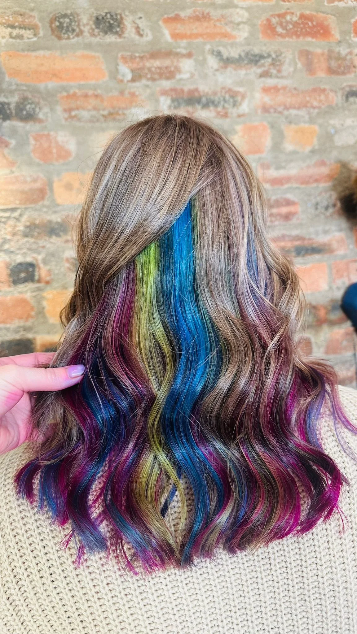 Back of a woman with long wavy hair dyed in rainbow colors including pink, yellow, green, blue, and purple, against a brick wall background.