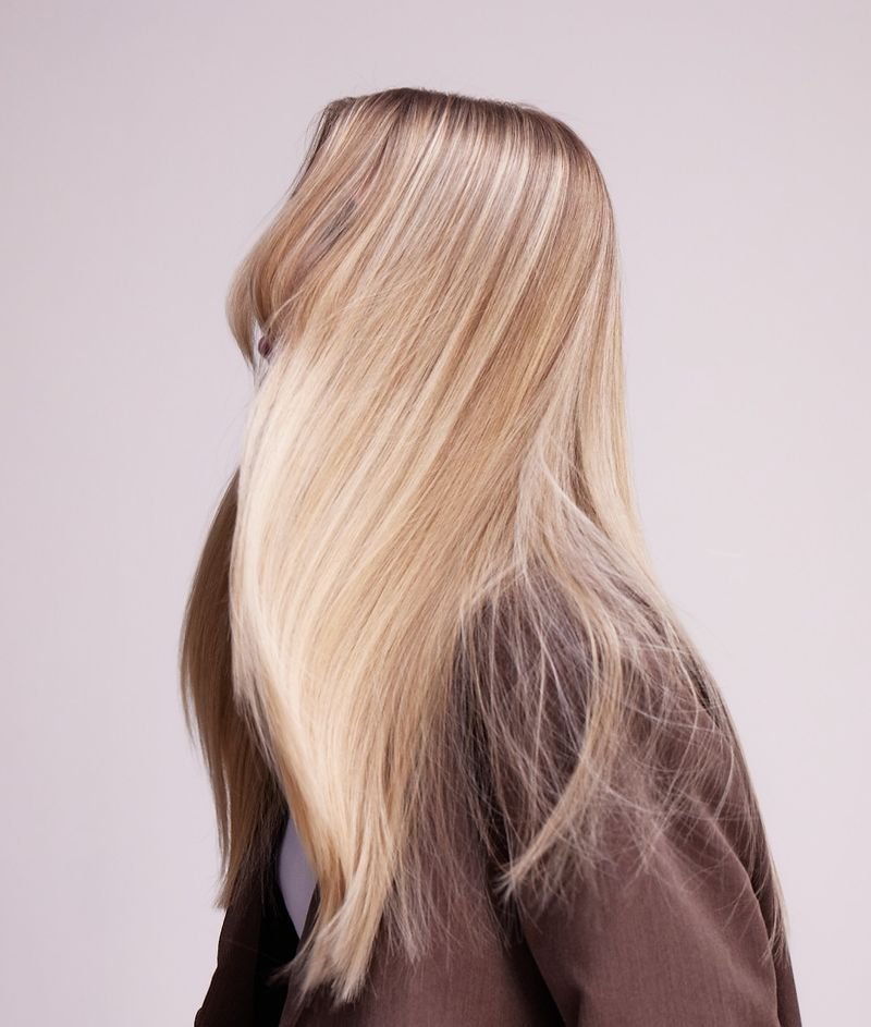 A person with long, straight, blonde hair covering their face, wearing a dark shirt or jacket against a plain light background.