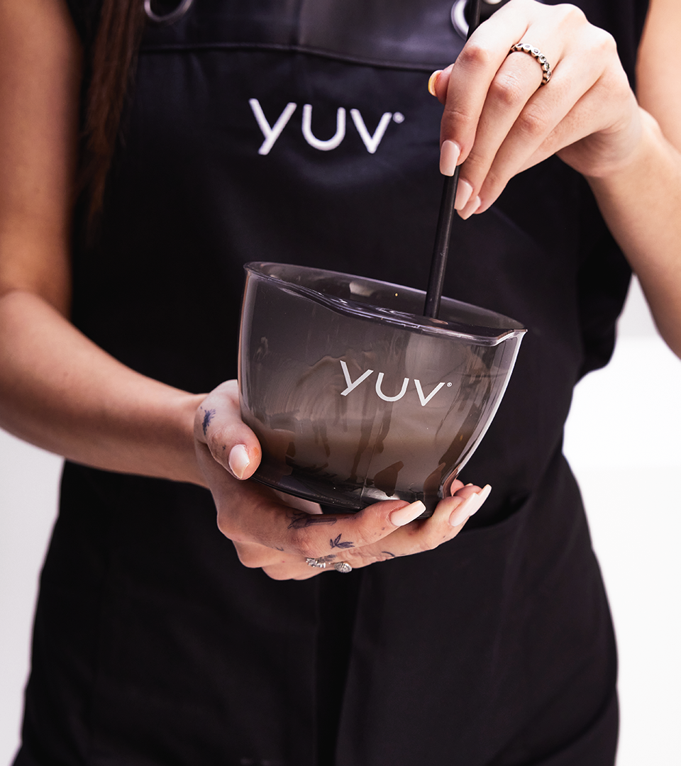 A woman holding a dark glass bowl and stirring it with a stick. She is wearing a black apron with the letters 'YUV' on it, and has rings on her fingers and tattoos on her hand.