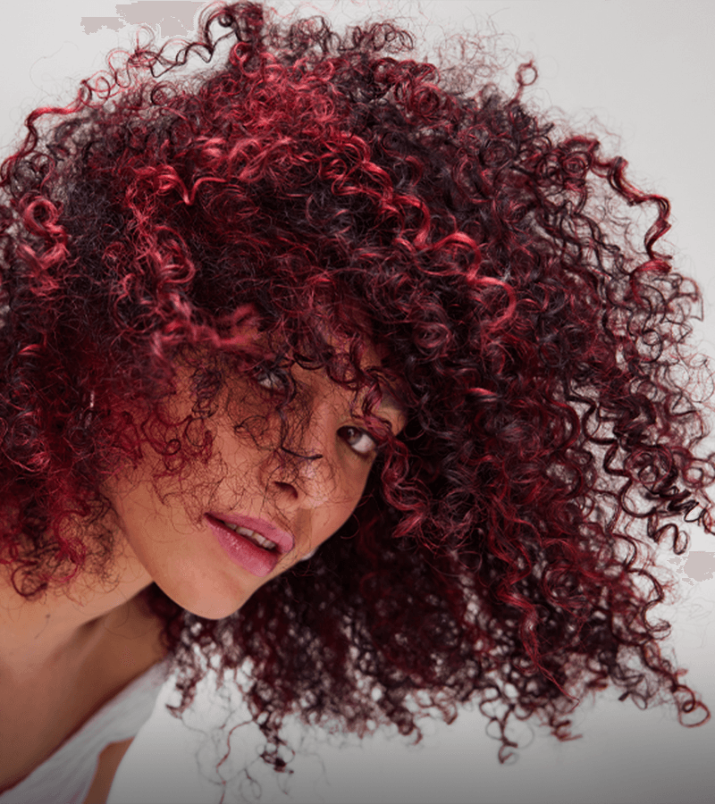 Close-up of a woman with wild, curly hair that is dark with vibrant red highlights, partially covering her face, with a neutral gray background.