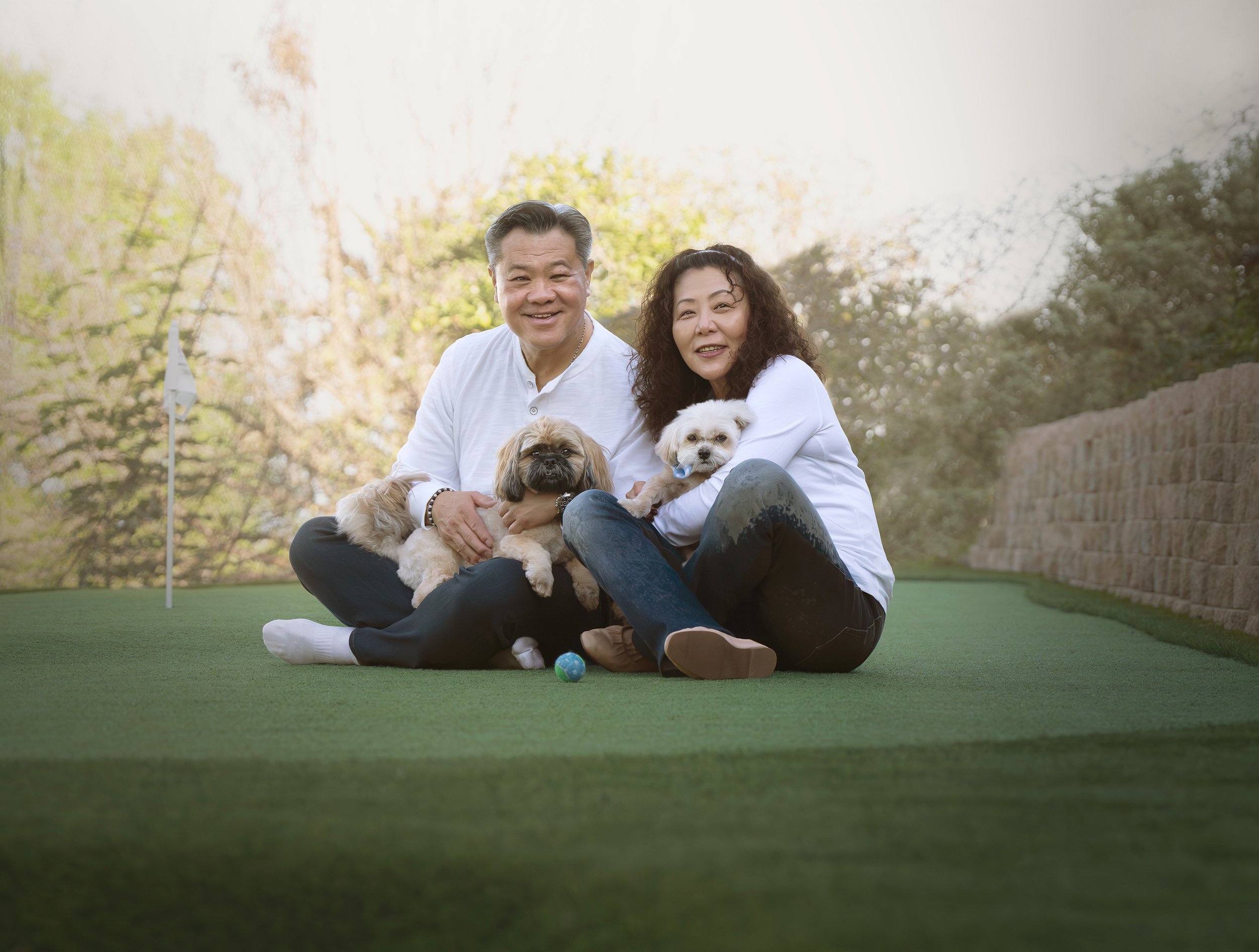 A happy middle-aged Asian man and woman sitting on a green outdoor mini golf putting green with two small dogs, smiling at the camera, surrounded by trees and a stone wall in the background.