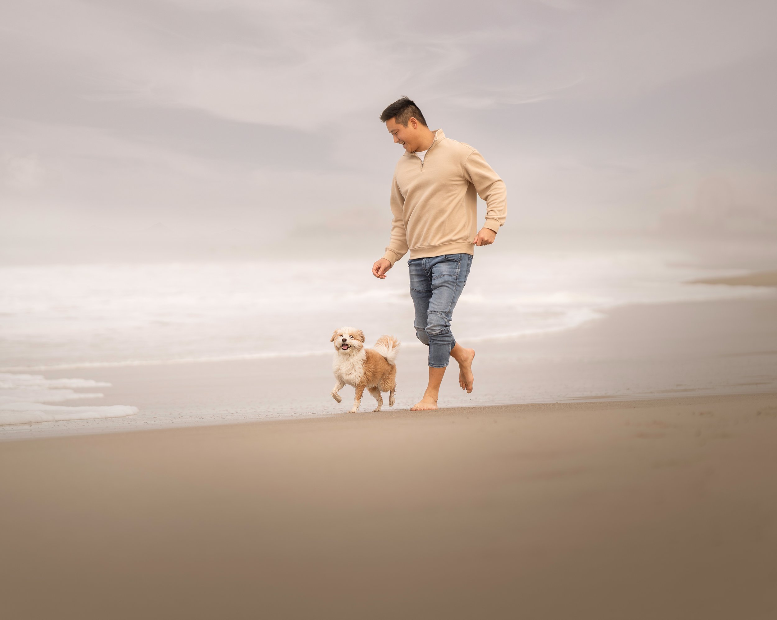 A man walking on the beach with a small, fluffy poodle mix dog. The sky is overcast and the ocean waves are visible in the background.