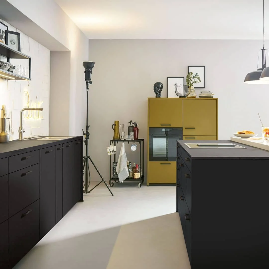 Modern kitchen with black lower cabinets, a yellow upper cabinet, a black hanging light fixture, and decorative items on top, including framed pictures, a bust, and plants.