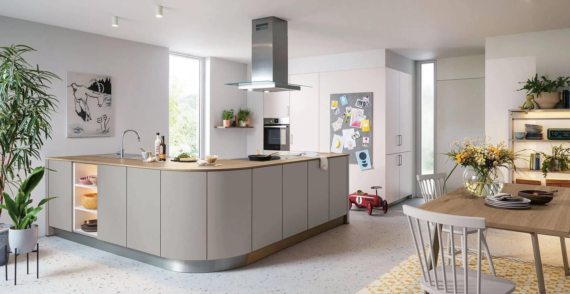 Modern kitchen with white cabinetry, wooden countertops, potted plants, a dining table with plates and silverware, and a red children's ride-on toy. There are large windows, a stainless steel range hood, and a bulletin board with photos and drawings.