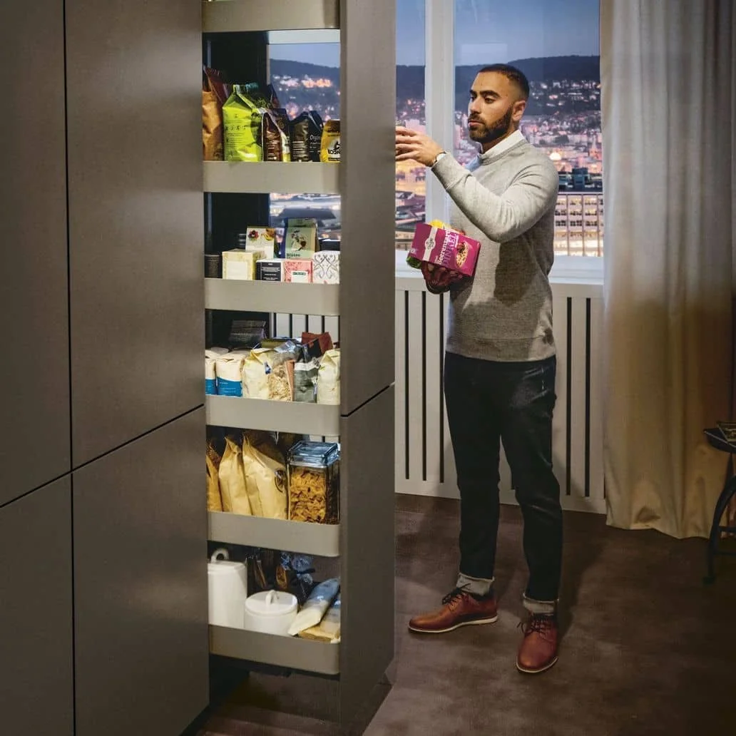 A man is standing in front of an open pantry, reaching for an item on the top shelf, holding a package in his left hand. The pantry contains various food and household items, and there is a window with a city skyline in the background.