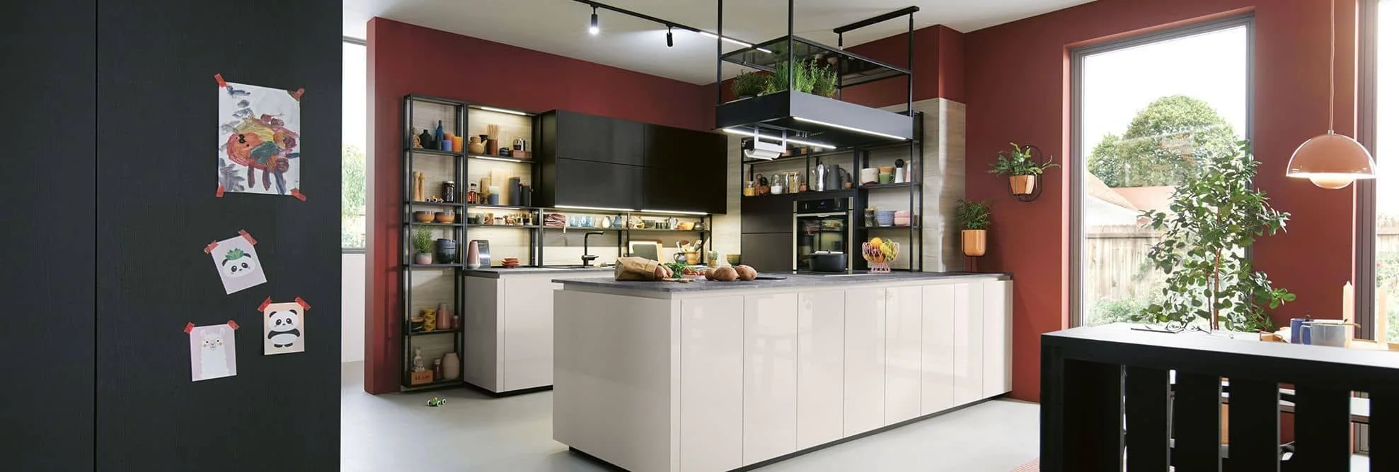 Modern kitchen with red accent walls, open shelving, white cabinets, and large window with outdoor view, decorated with plants and artwork.