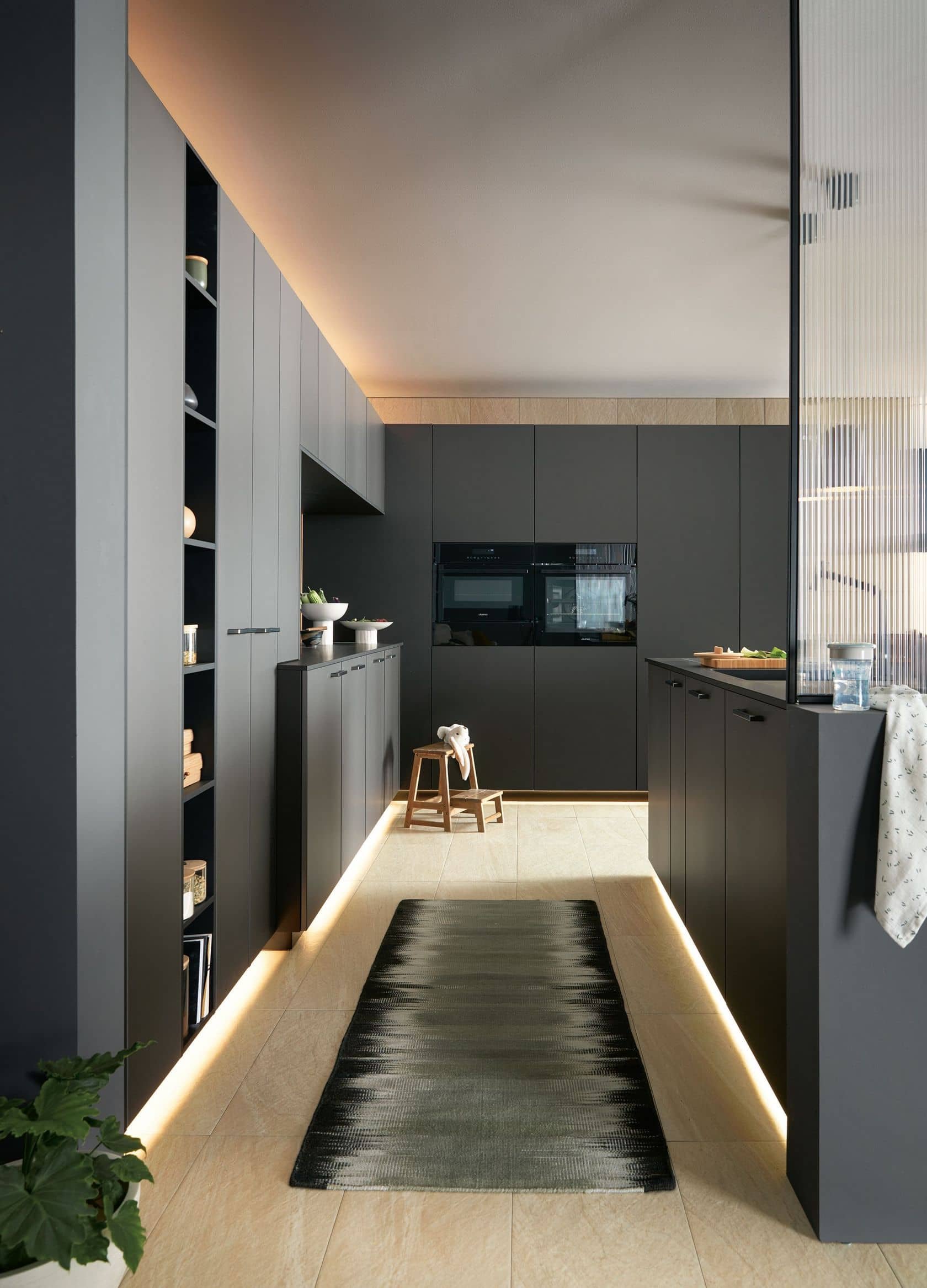 Modern kitchen with dark gray cabinets, beige tile flooring, black accents, and under-cabinet lighting. Small wooden stool with a doll on it, a black runner rug, and some decorative items on the countertops.
