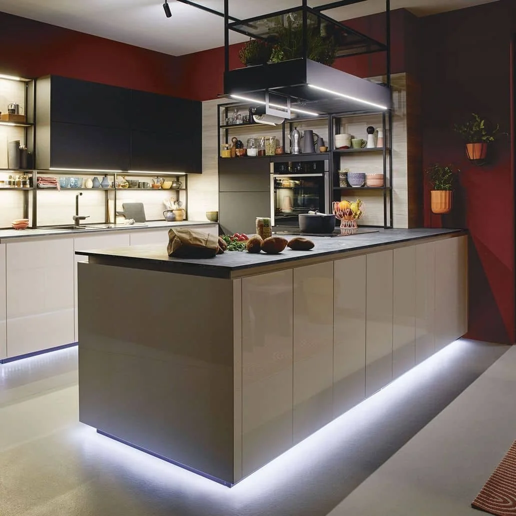 Modern kitchen with black, beige, and red accents, featuring an island with under-cabinet lighting, open shelving with kitchenware, and potted plants.