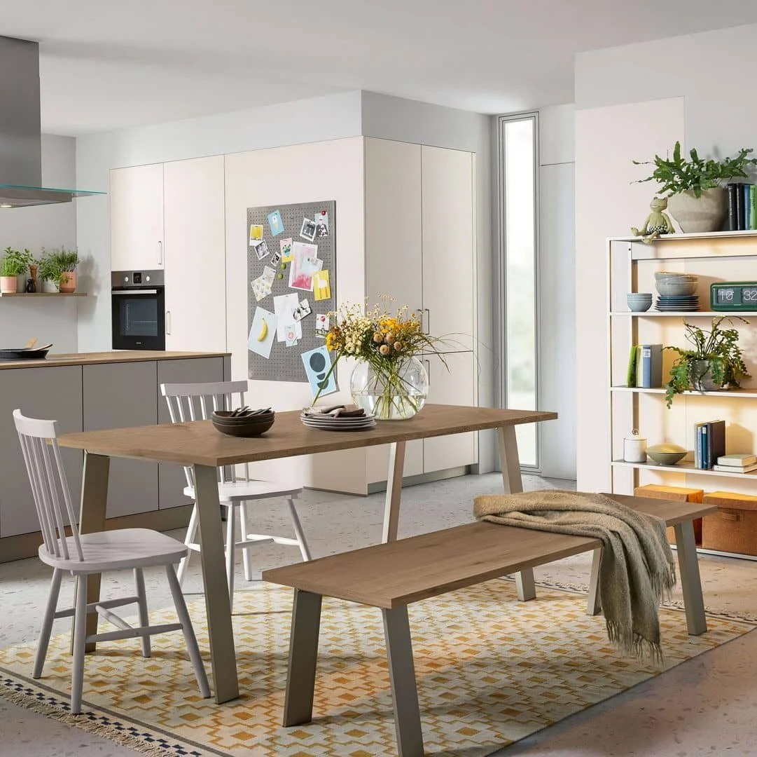 Modern kitchen and dining area with a wooden table, white chairs, a bench with a blanket, a vase of flowers, shelves with plants and books, and light-colored cabinetry near a large window.
