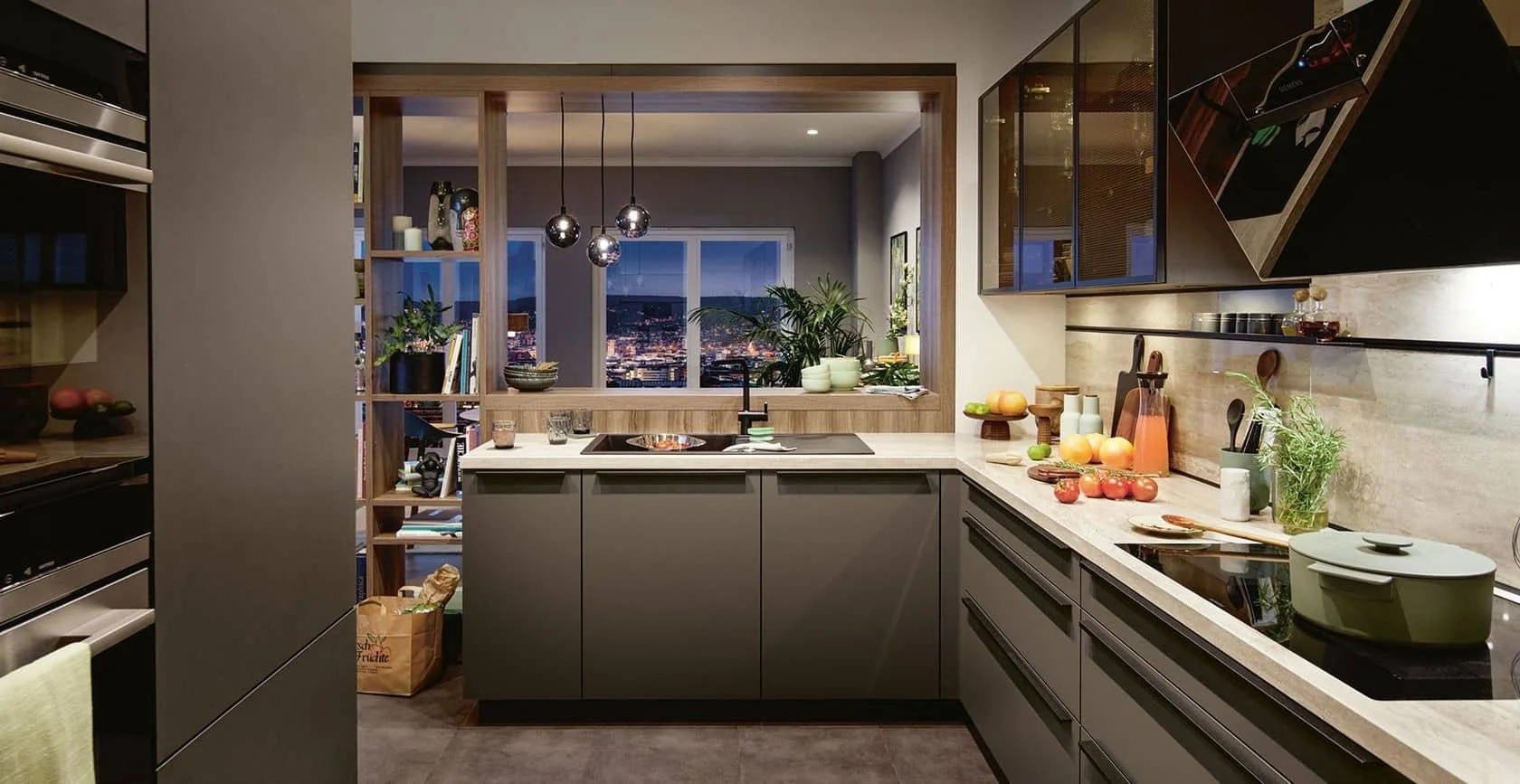 Modern kitchen with gray cabinets, marble countertops, and a city view through a window, decorated with fruit, plants, and kitchen utensils.