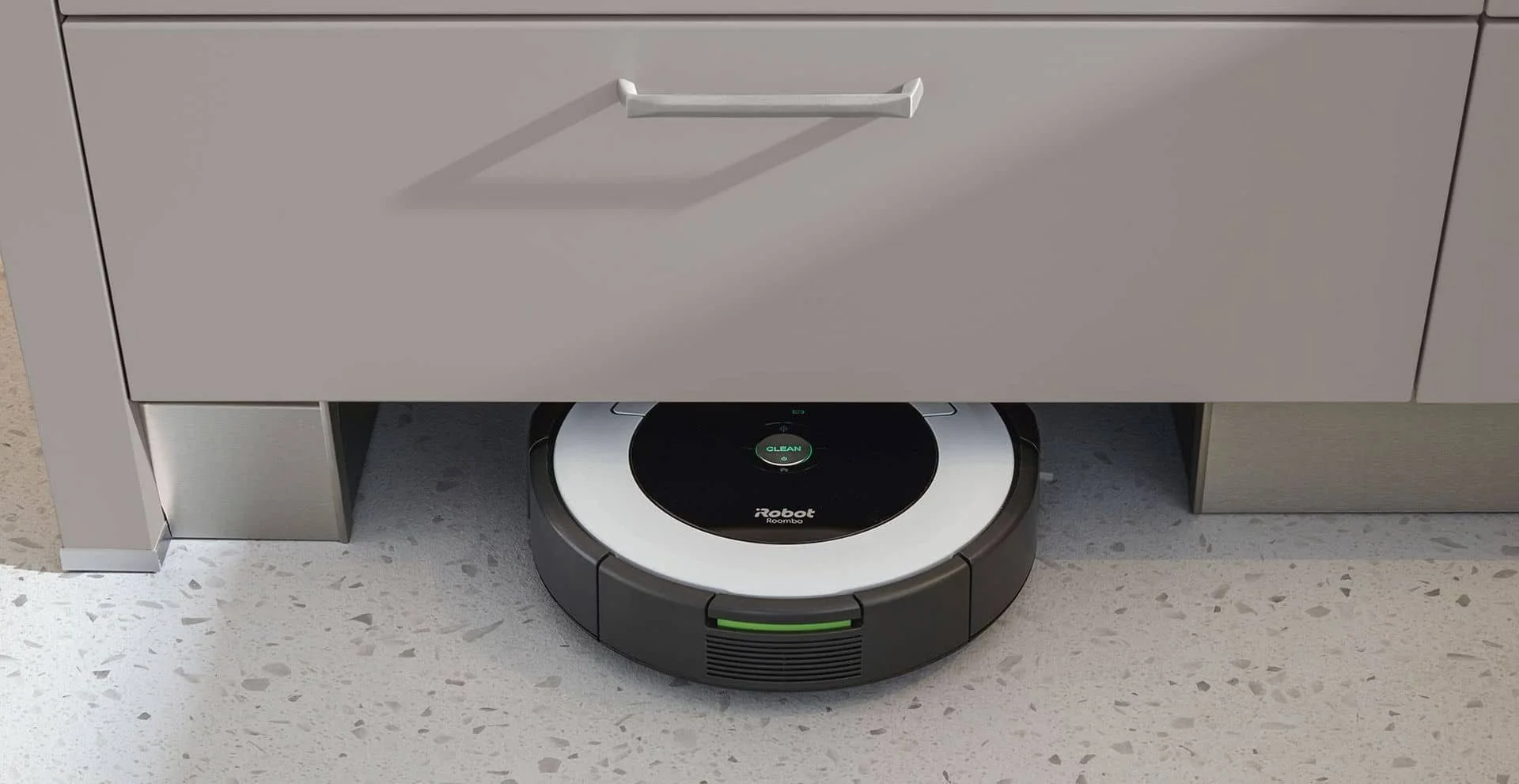 A robotic vacuum cleaner under a kitchen drawer on a light-colored speckled floor.