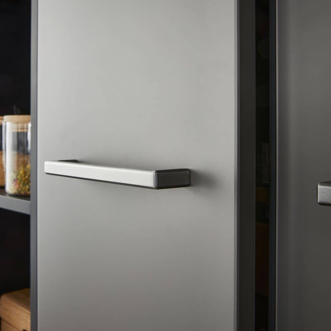 Close-up of a gray kitchen refrigerator door with a silver handle, partially open to reveal shelves with jars of spices and food items.