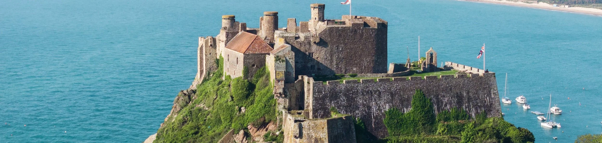 A historic stone castle on a hill overlooking the water, with boats docked nearby and flags flying.