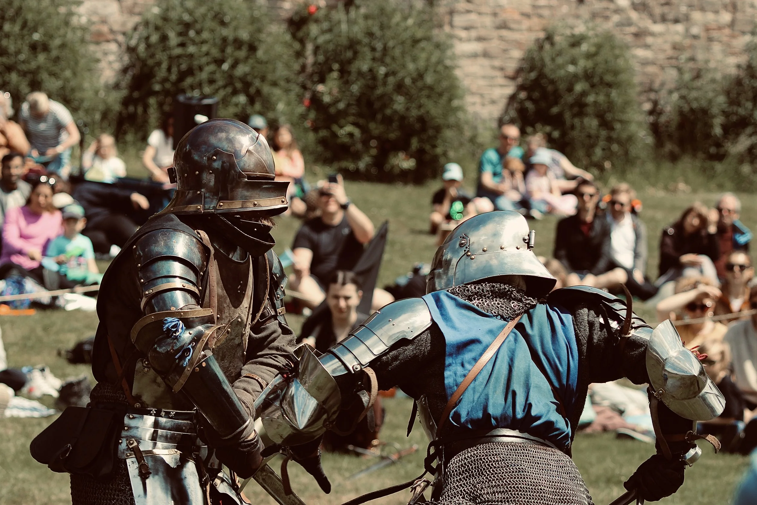 Two medieval knights in armor duel with swords during a reenactment event in front of an audience seated on grass.