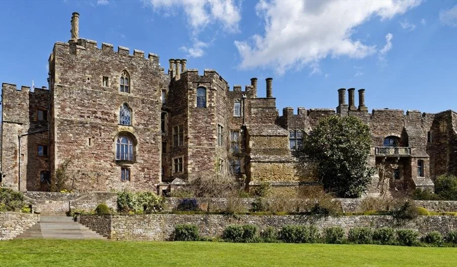 An old stone castle with multiple towers and battlements, surrounded by a manicured lawn and gardens, under a partly cloudy sky.