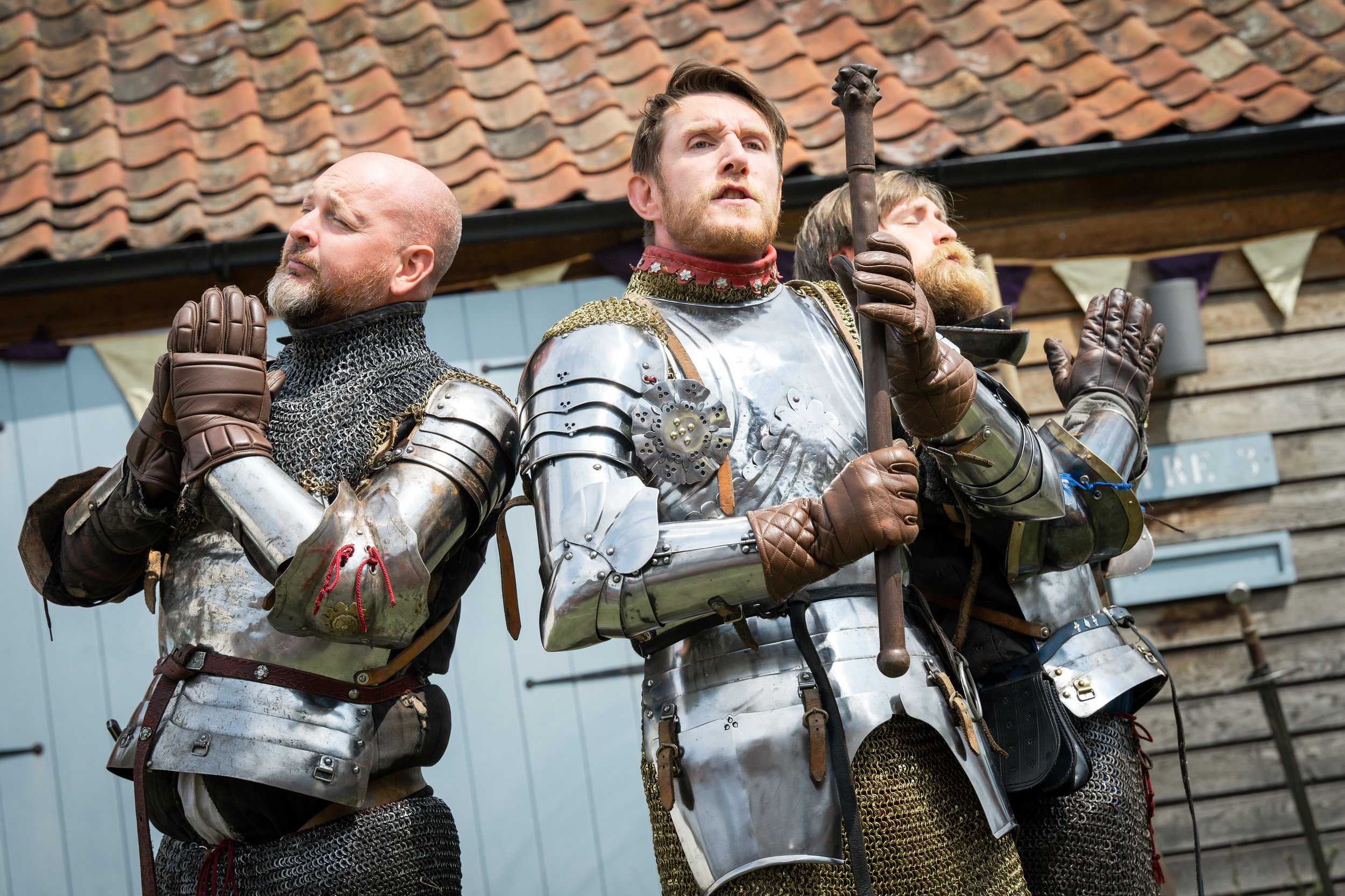 Three men dressed in medieval armor standing with their eyes closed and hands in a prayer-like gesture, outdoors, with a wooden building and a tiled roof in the background.