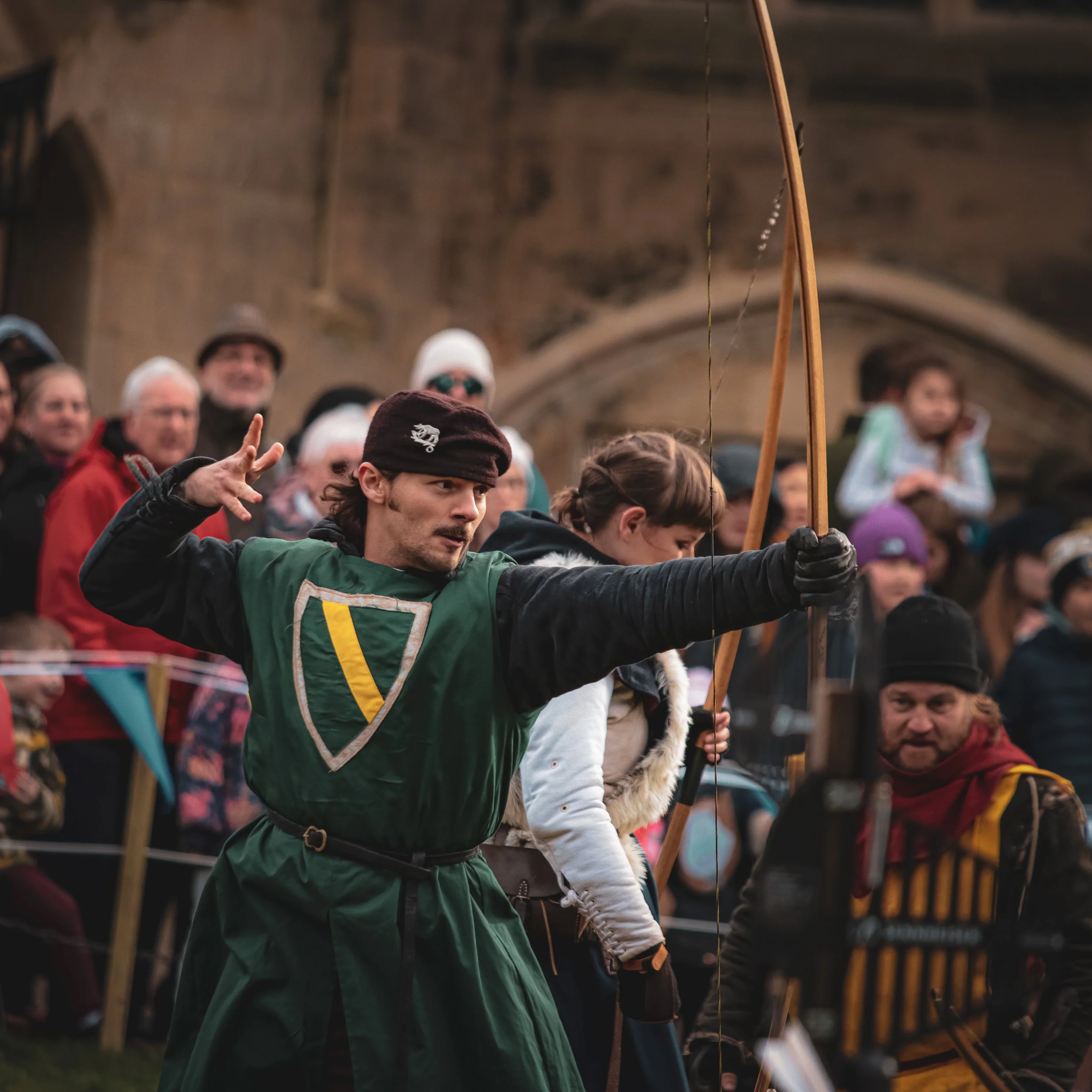 A man dressed as a medieval archer aiming with a bow in a reenactment, surrounded by spectators.