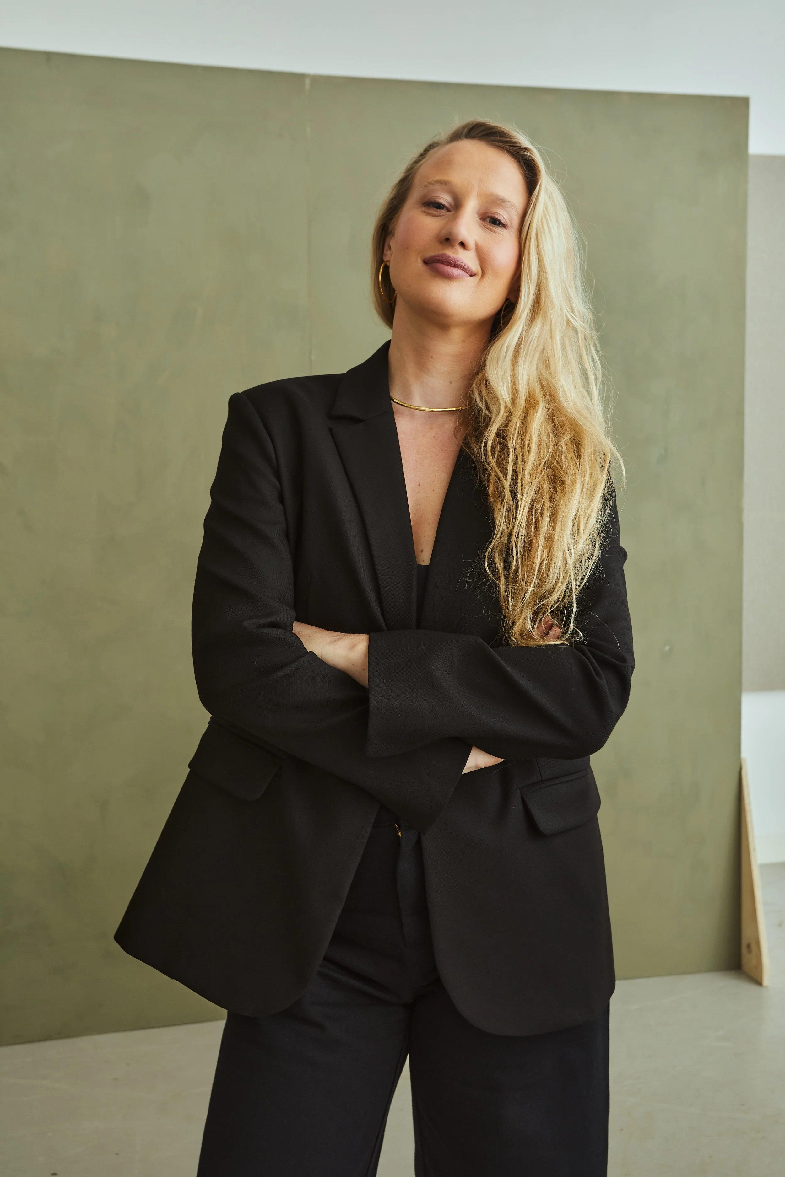 A woman with long blonde hair wearing a black blazer and pants standing with arms crossed in front of a green and gray background.