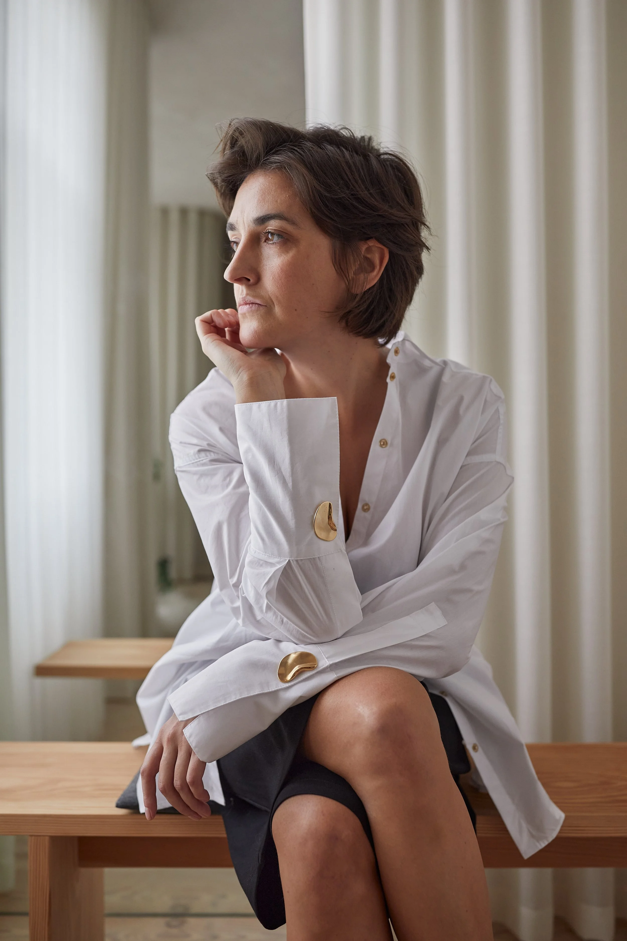 A woman with short brown hair sitting on a wooden bench in a room with cream-colored curtains, wearing a white button-up blouse and shorts, looking thoughtful and gazing out of a window.