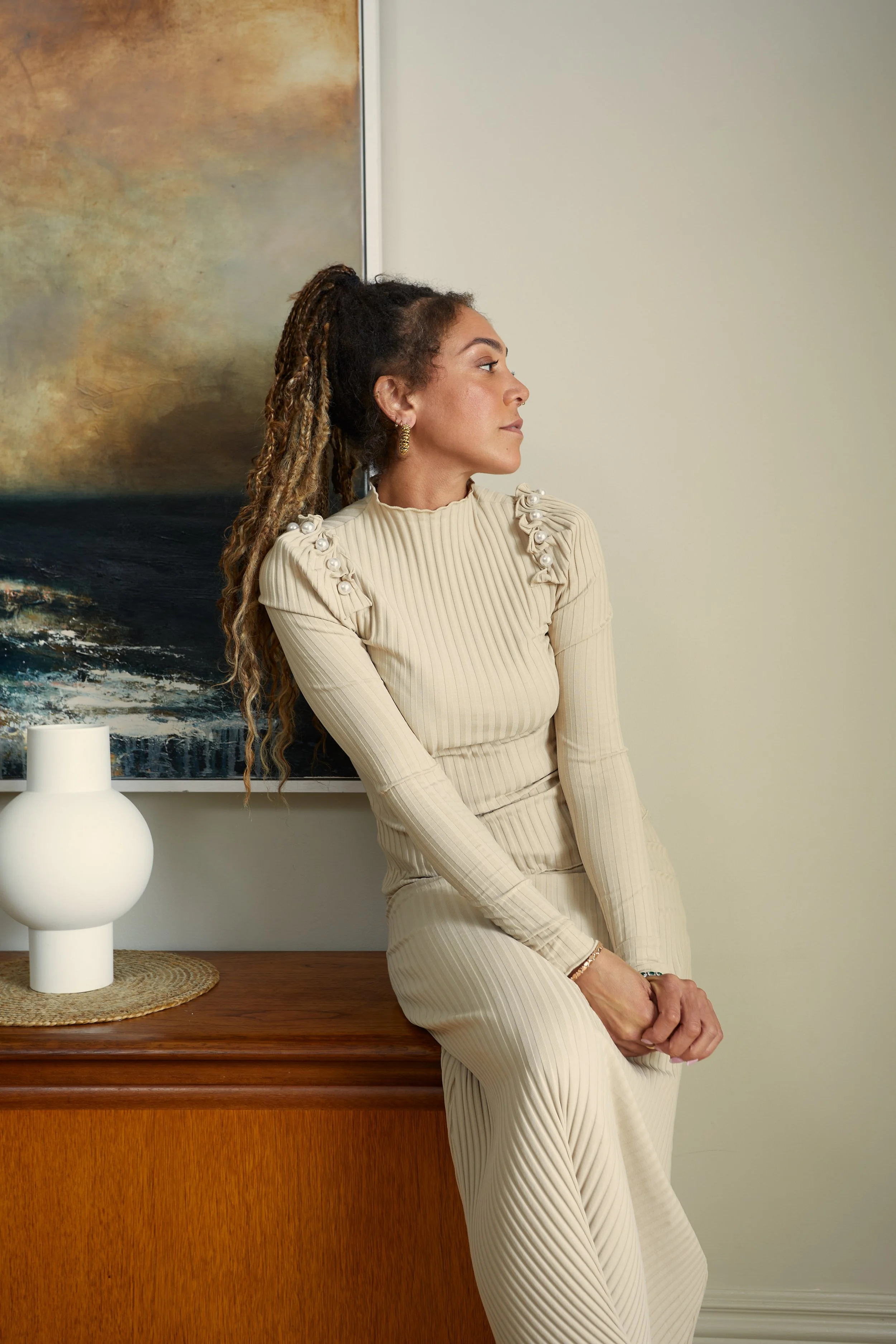 A woman with dreadlocks leaning against a wooden table, wearing a cream-colored ribbed dress with pearl embellishments on the shoulders, in a room with abstract art on the wall and a white ceramic vase.
