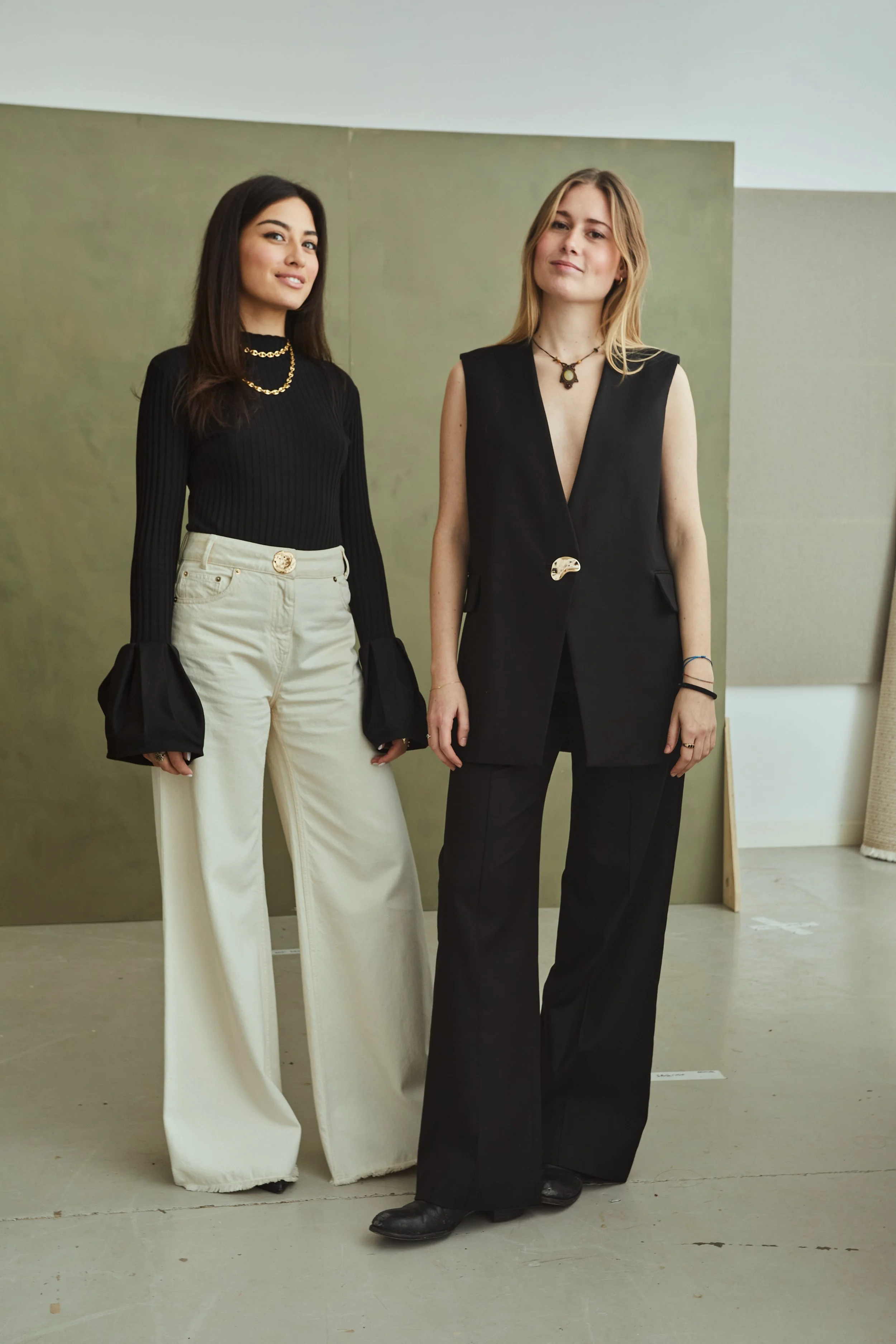 Two women standing side by side in fashionable outfits, one in a black long-sleeve top and cream wide-leg pants, the other in a black sleeveless blazer and matching pants.