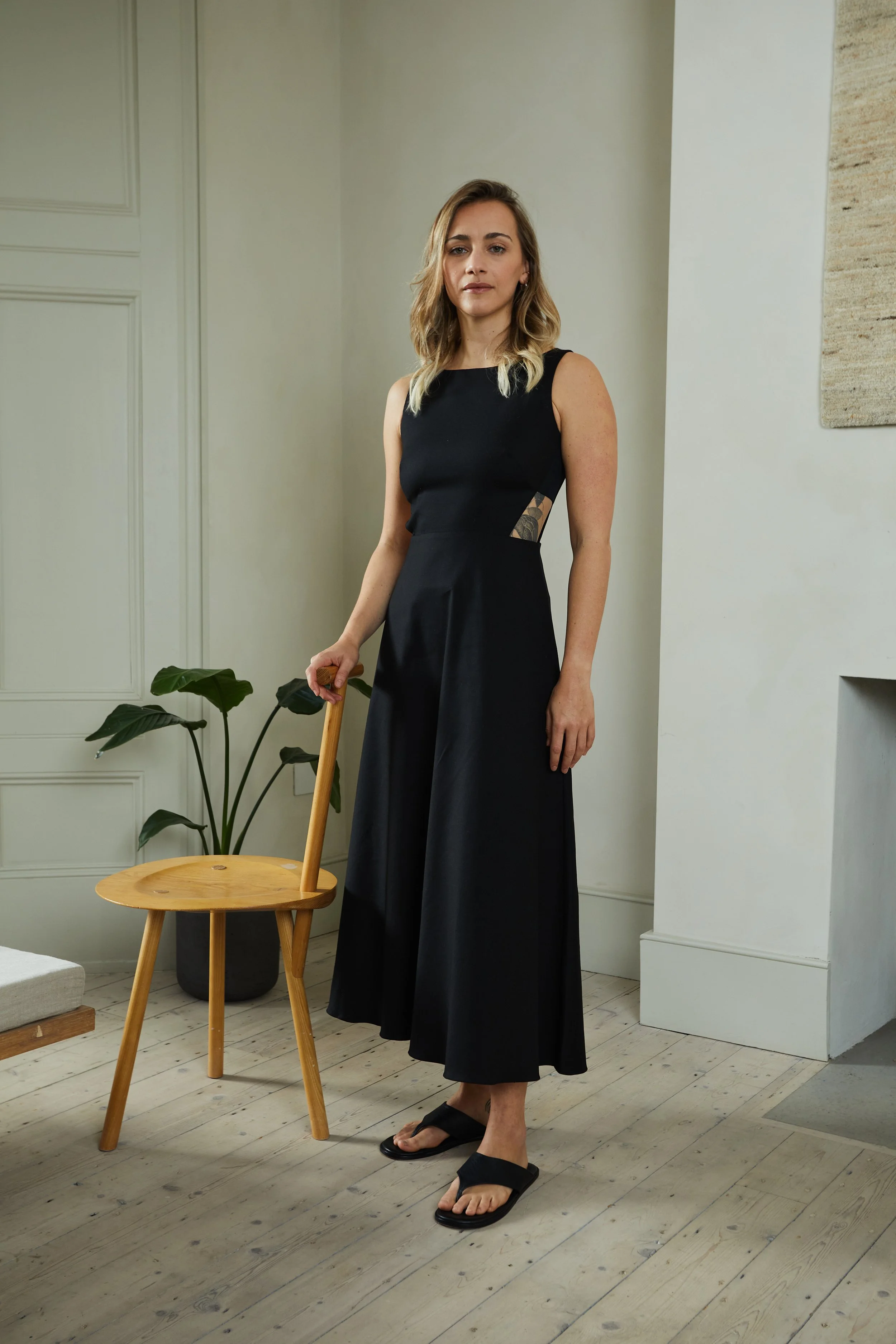 Woman in a black sleeveless dress and sandals standing inside a room with wooden flooring, holding a wooden chair, with a potted plant and white walls in the background.