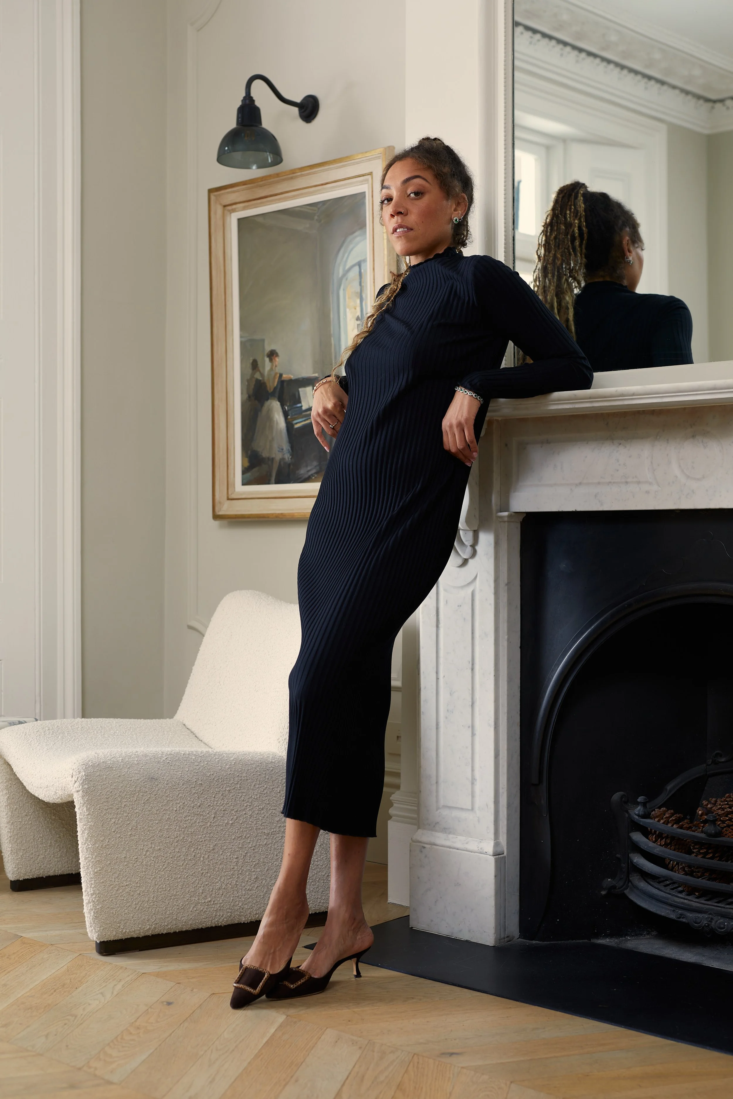 A woman in a long dark dress and heels leaning against a fireplace mantel in a living room, with a framed painting and a mirror behind her.