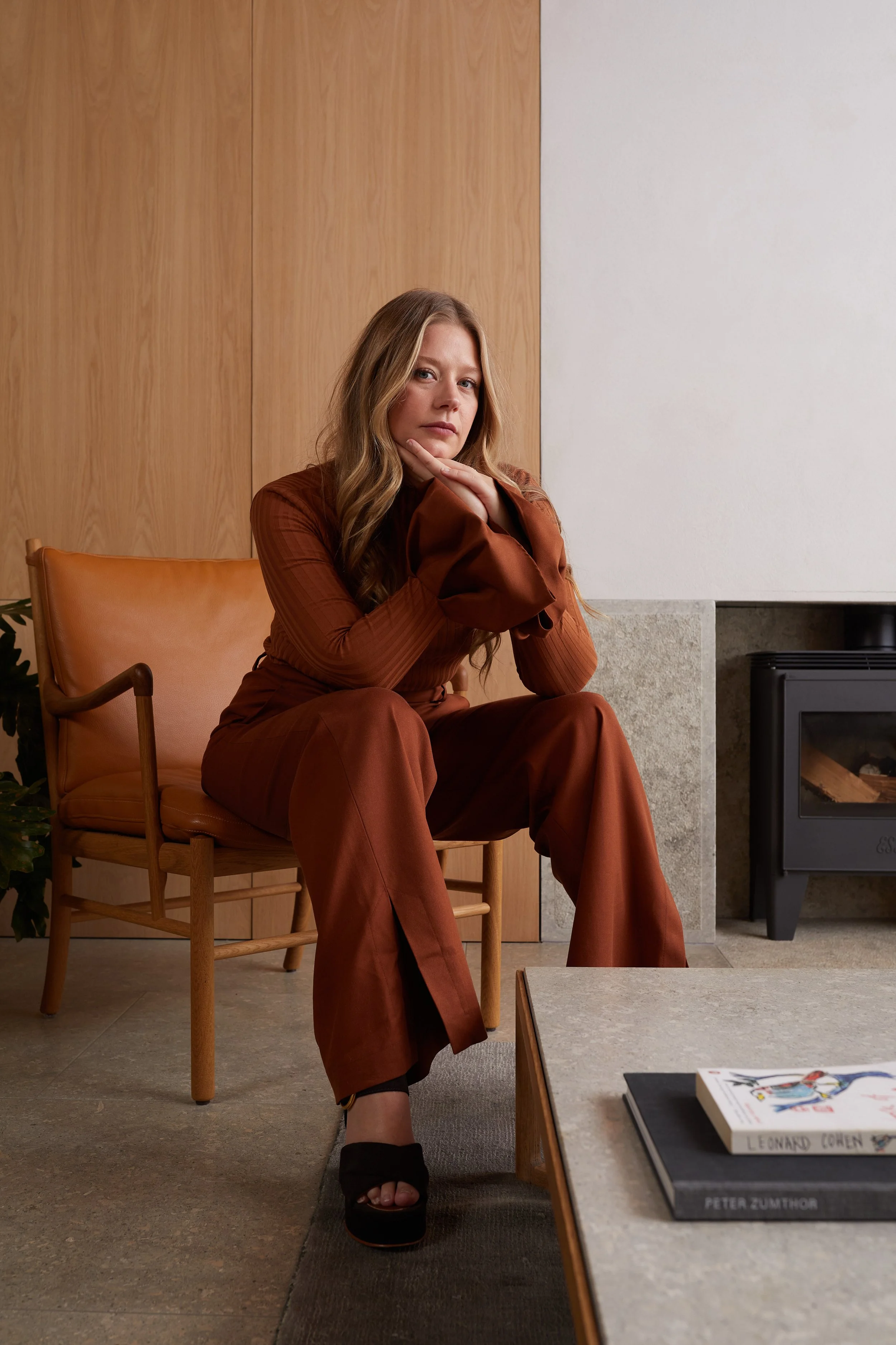 A woman with long blonde hair is sitting on a wooden armchair with leather cushions in a modern living room, wearing a matching brown outfit, looking at the camera with a thoughtful expression.