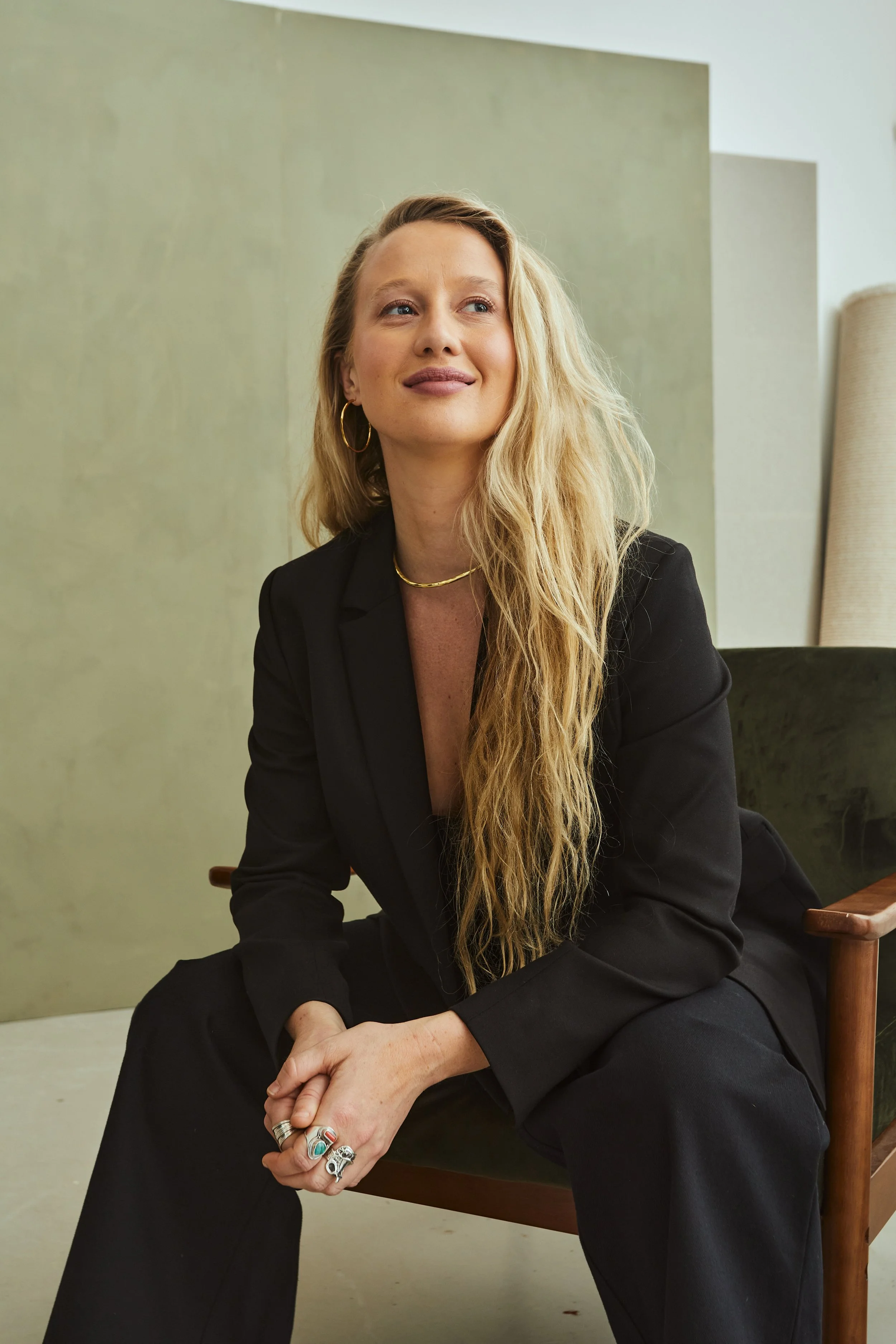 A woman with long blonde hair, wearing a black suit, gold hoop earrings, a gold necklace, and multiple rings, sitting on a green velvet armchair in an indoor setting with muted green and beige background.