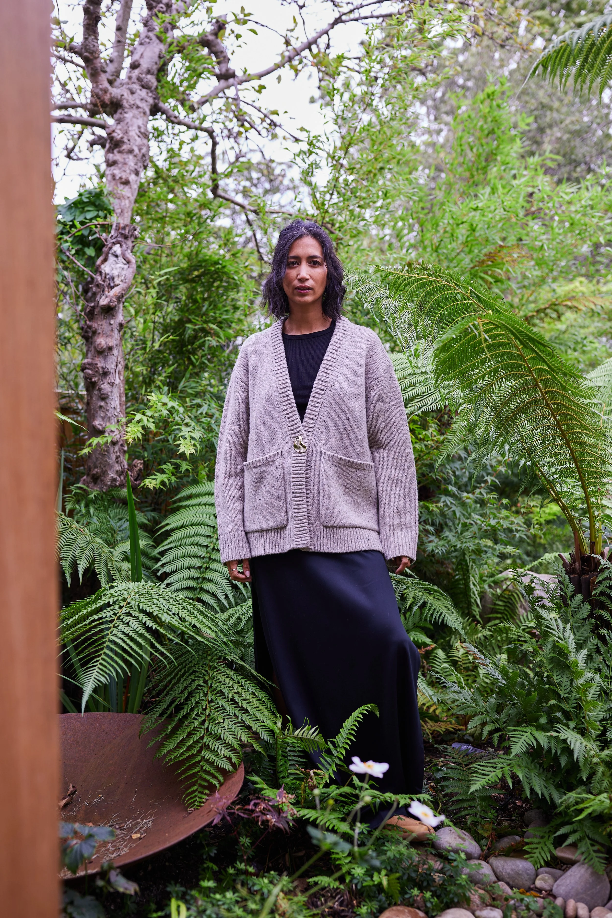 A woman standing in a lush, green garden with dense foliage and ferns around her, wearing a gray cardigan over a black dress.
