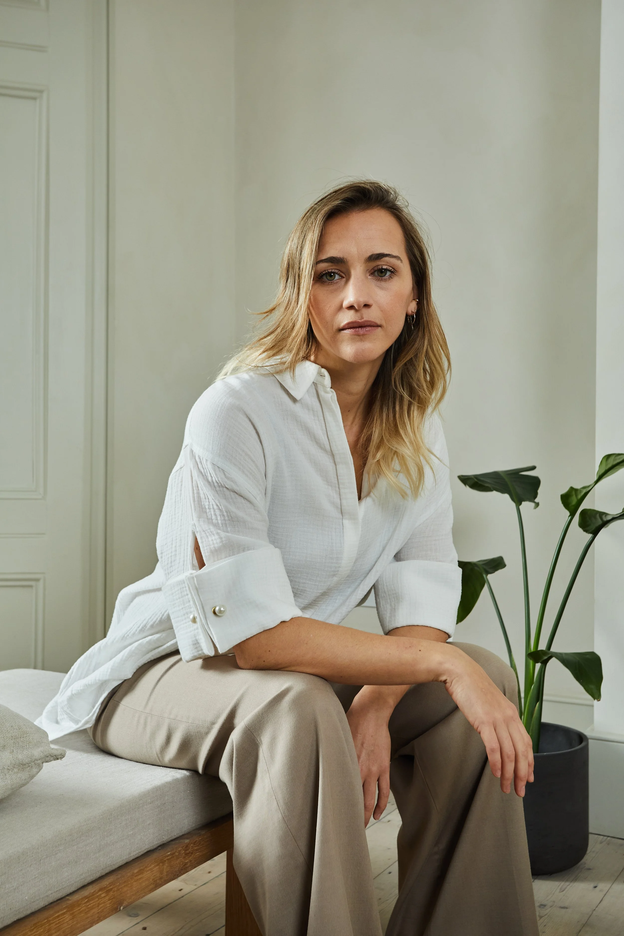 A woman with blonde hair wearing a white blouse and beige pants sitting on a sofa next to a potted plant in a room with neutral decor.