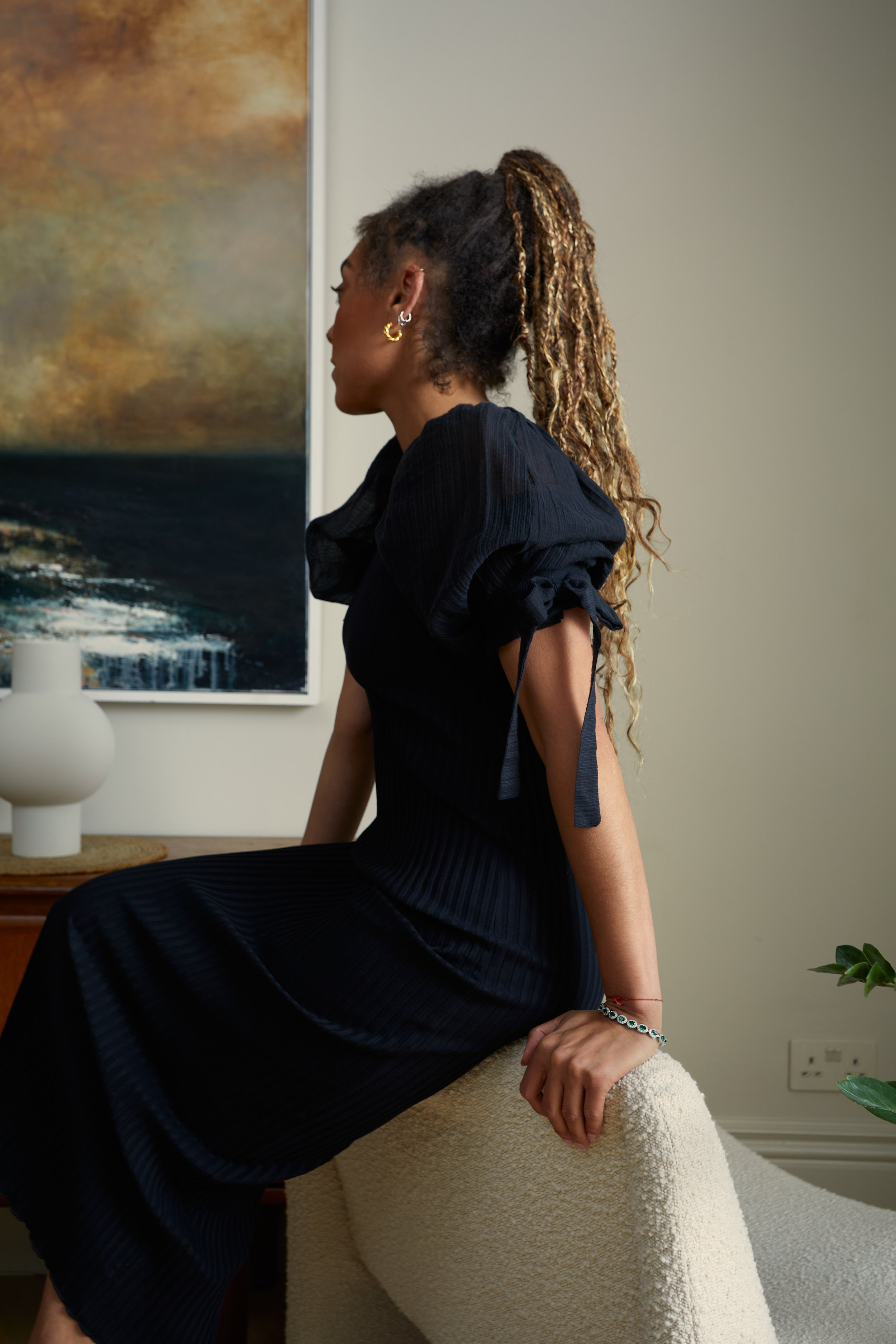 A woman with curly hair styled in a high ponytail with blonde highlights, wearing a black dress with puffed sleeves, sitting on a cream-colored chair in a room with a painting, a white decorative vase, and a plant.