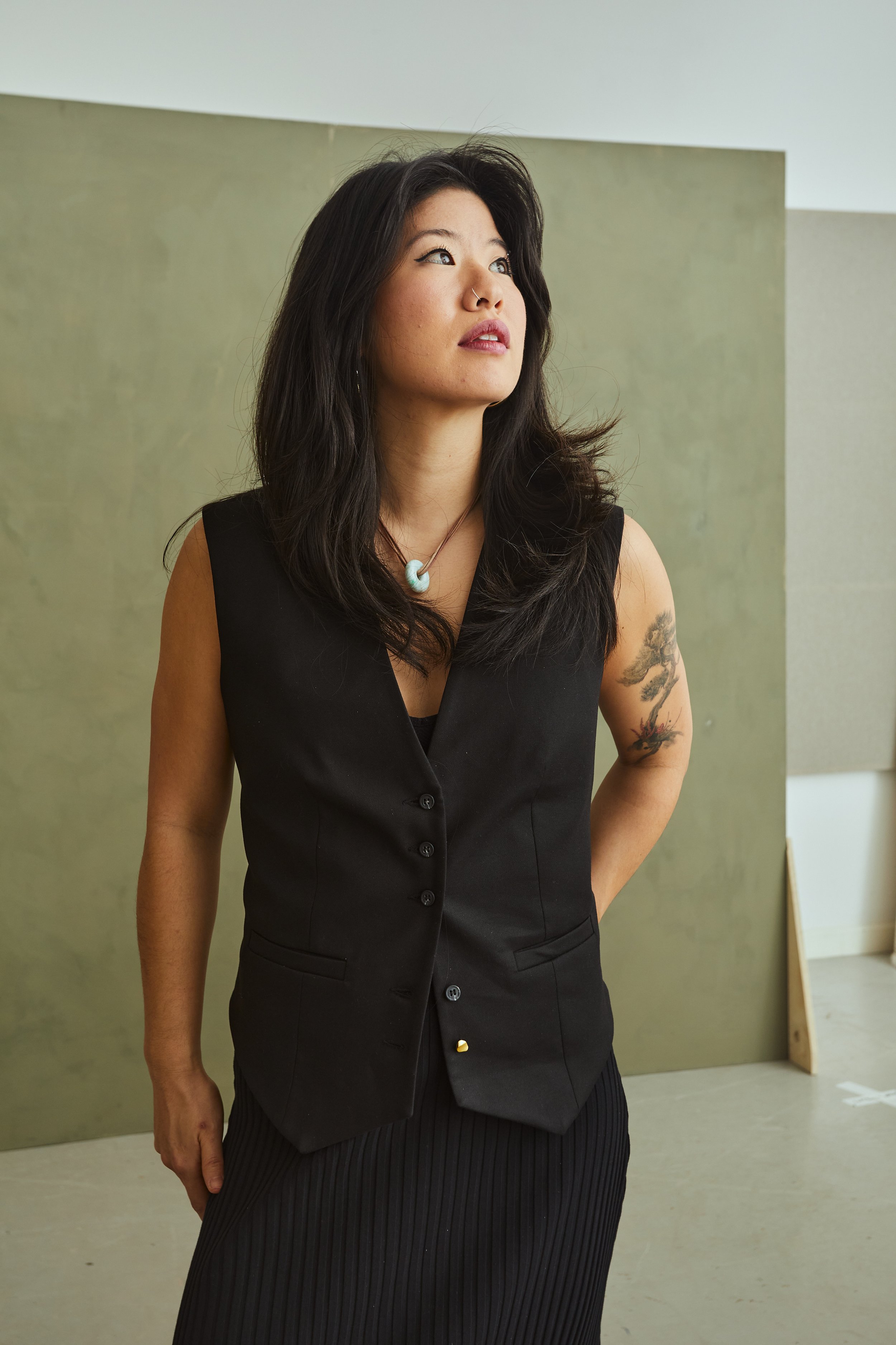 A woman with black hair, wearing a sleeveless black vest, standing indoors with a green and gray background, looking to her left.
